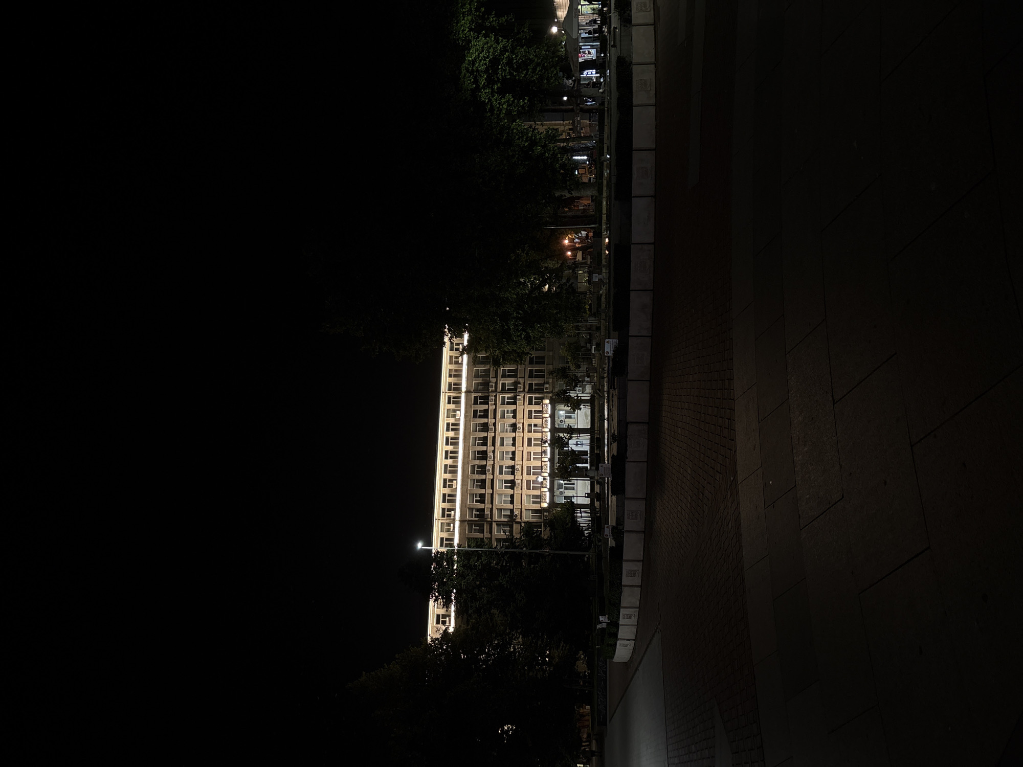 This image depicts a nighttime scene in a public square in Varna, Bulgaria. A multi-story building, approximately five floors high, dominates the background. It features a light-colored facade with numerous rectangular windows, many equipped with small balconies or ledges and external air conditioning units. The building's top floor and roofline are illuminated by a continuous line of warm white light, while several windows on lower floors show interior illumination. A tall street light with a bright white lamp stands in front of the building, slightly to the left of the center.
The midground and foreground comprise a paved public square with varying patterns of stone tiles and blocks. A low, curved or angled concrete structure, possibly a planter or fountain, defines an area within the square, and along its visible side, multiple rectangular plaques or relief designs are embedded. Dense, dark trees line the left and right perimeters of the square, their foliage partially visible against the night sky and minimal ambient light. A cluster of smaller trees and bushes occupies the central square area. On the far right, behind some trees, an illuminated outdoor kiosk or café is visible, where several indistinct figures can be observed seated or standing, engaged in unspecified leisure activities. No other individuals are clearly discernible in the broader square. The overall environment is an urban setting, dimly lit by artificial sources.