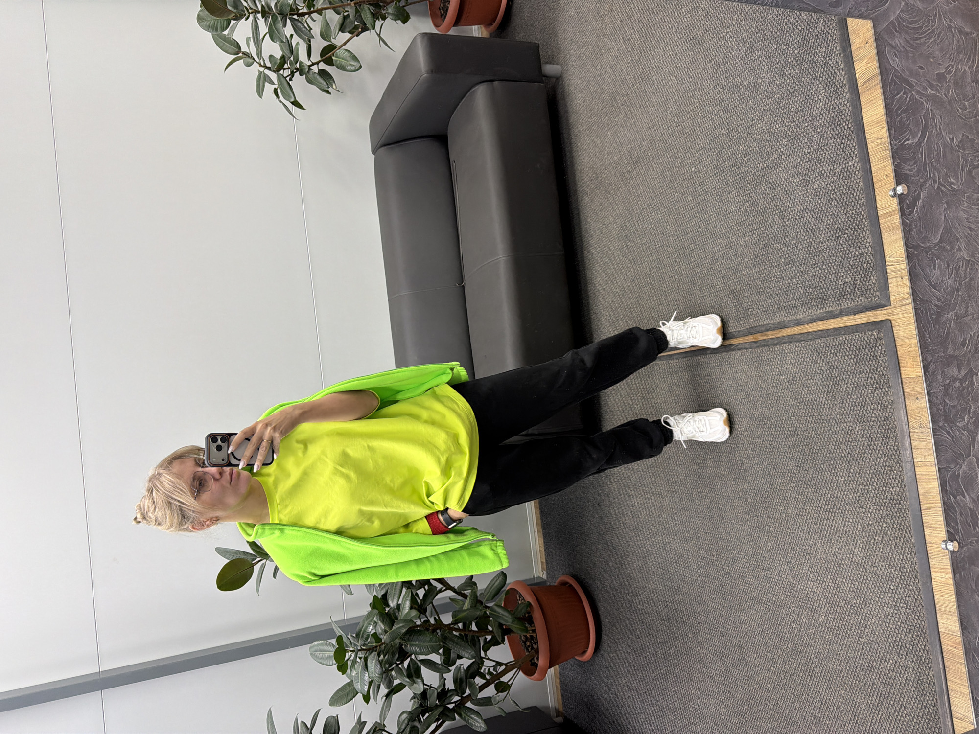 A blonde woman in glasses is taking a selfie in what appears to be an office setting in Chaikovsky, Russia. She is wearing black pants, white sneakers, and a bright yellow shirt with a matching green hoodie. She is standing on a grey carpet between two potted plants with shiny green leaves. Behind her is a black leather loveseat, and a plain white wall. The carpet has a dark wooden trim along its edges. The woman is holding a phone in her left hand. The scene shows her directly. Part of the building's architectural plan is evident, displaying its materials and design.