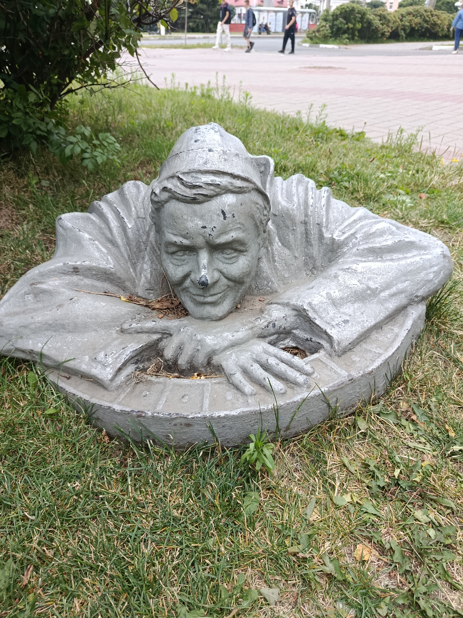 The image shows a concrete sculpture of a man's head and upper torso emerging from the ground. The sculpture is gray and appears weathered, with some black paint chips. The man has a smiling expression and is wearing a cap and a garment with a ruffled collar. His hands are resting on a circular, textured base that is partially embedded in the grass. The sculpture is located on a grassy area next to a paved walkway, with a street and buildings visible in the background. There are a few people walking in the background, and some green bushes and trees to the left of the sculpture. The overall environment suggests an outdoor public space, possibly a park or a plaza in the city of Zhlobin, Belarus.