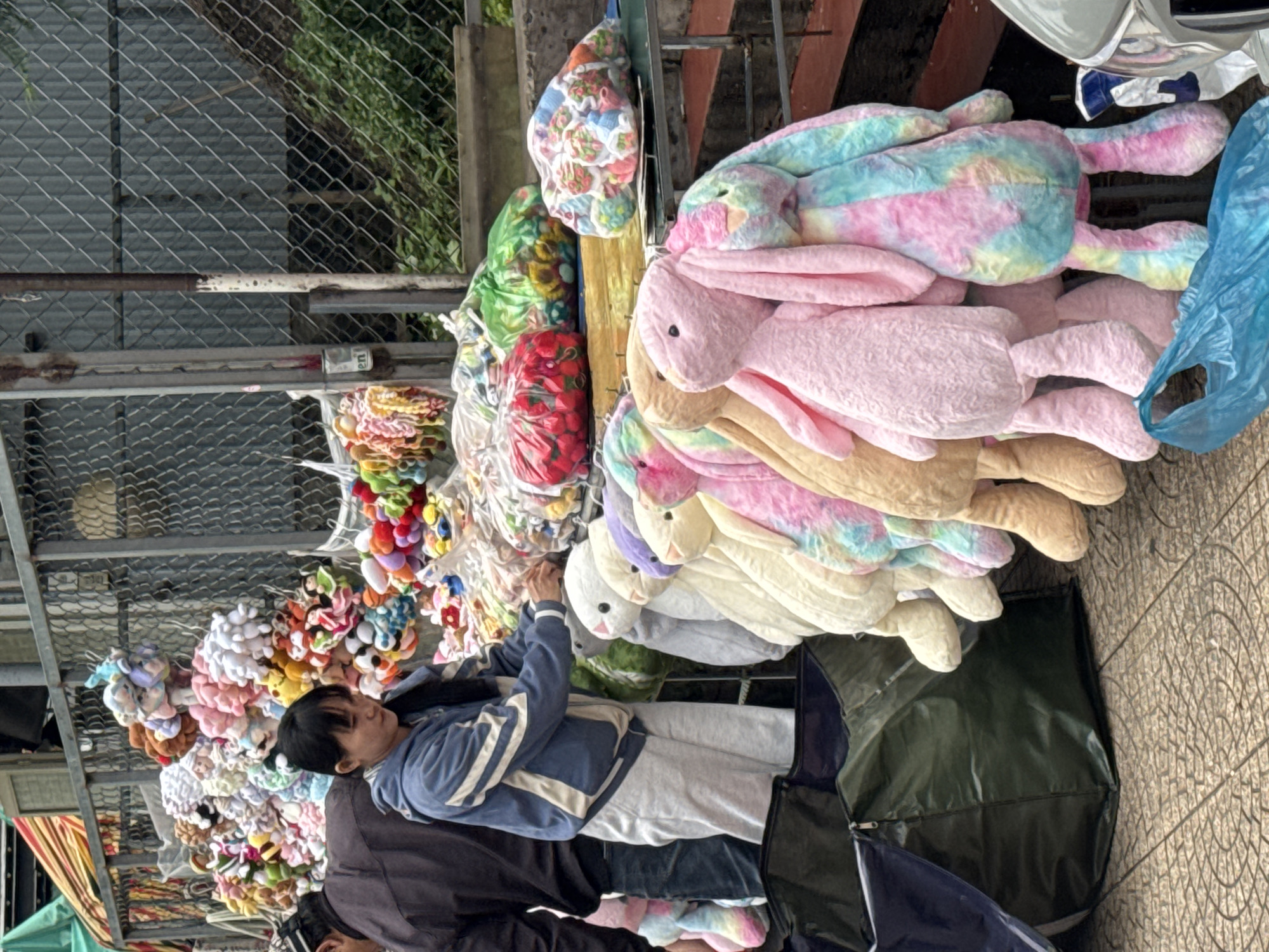This image captures a street vendor stall in Da Lat, Vietnam, specializing in plush toys and small items. In the foreground-center, a woman with dark hair, wearing a blue hooded jacket with white stripes and light gray sweatpants, reaches toward merchandise displayed on a table. To her left, a man wearing a dark jacket and a cap is partially visible from the back.
The stall features a large array of stuffed animals. In the mid-ground, a prominent display includes several large, long-eared plush bunnies hanging vertically. These bunnies vary in color, including solid pink, beige, white, purple, and a multi-colored tie-dye pattern. Behind the individuals, a dense collection of smaller stuffed animals and trinkets is arranged on various surfaces, with many items bundled in clear plastic bags. On a metal table towards the right, three larger plastic bags of merchandise are visible: one red bag containing numerous red, possibly strawberry-shaped, items; one green bag filled with various colorful objects; and one clear bag containing small, patterned items.
The ground surface is paved with light-colored interlocking concrete tiles featuring a circular design. Two large, dark green soft-sided storage bags or duffels are positioned on the ground on the left. A light blue plastic bag lies on the ground in the lower right corner. The background consists of a tall chain-link fence, behind which a sloped area with green vegetation is evident, along with a section of corrugated metal fencing. A part of a yellow and red striped awning is visible in the upper left corner, and a grey vehicle bumper is present on the far right.
