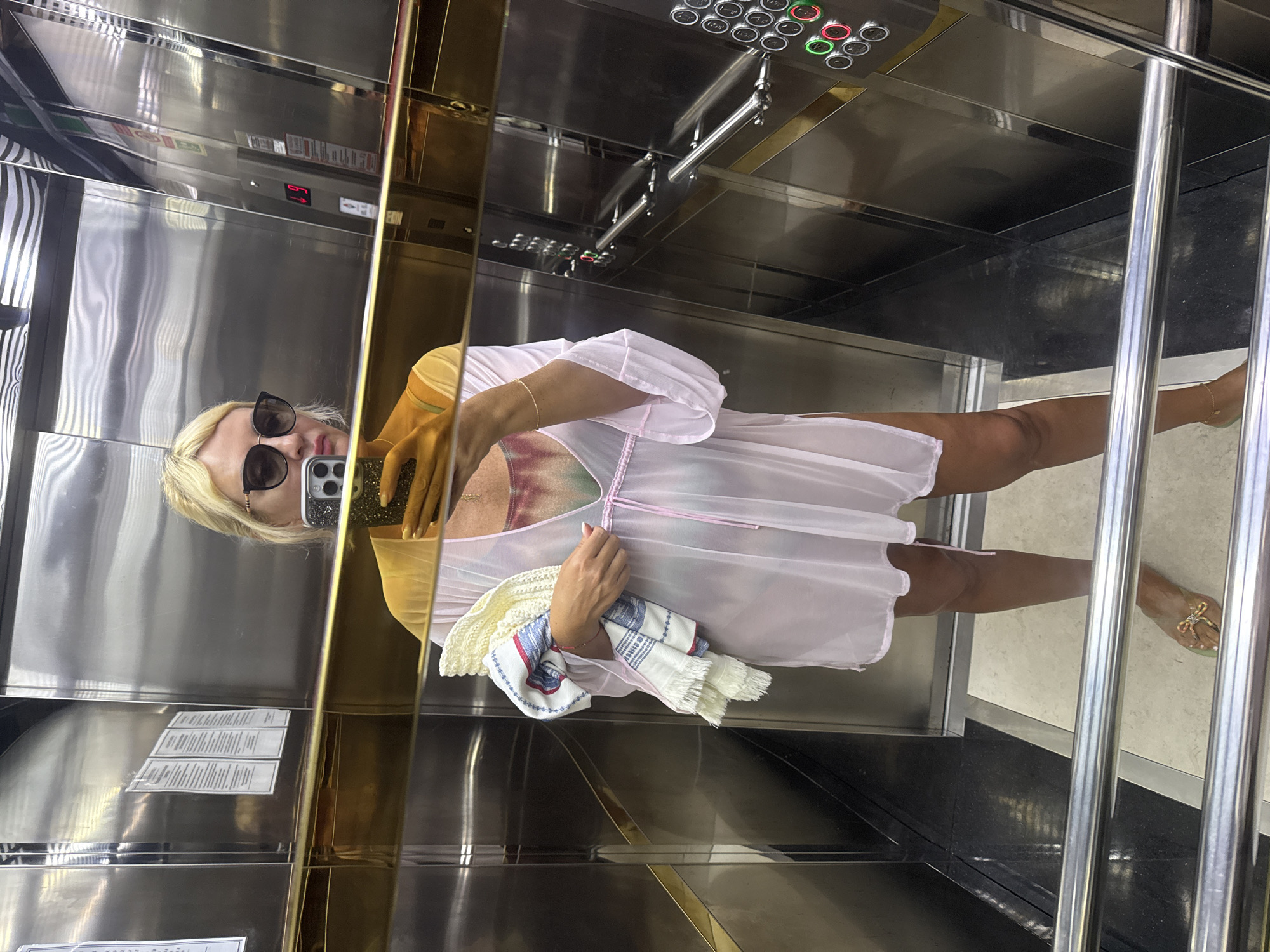The image shows a blonde woman in an elevator. She is wearing large, dark sunglasses and a light pink sheer cover-up over a tie-dye bikini top. She is holding a folded towel in her left arm. Her right hand is holding a smartphone, which is reflecting in the mirror. The elevator walls are made of polished stainless steel, with a gold-colored trim. There are elevator buttons visible on the right side of the image. The woman is wearing gold sandals. The overall impression is that the woman is dressed for a beach or pool outing, and the elevator is likely in a hotel or resort in Alanya, Turkey.