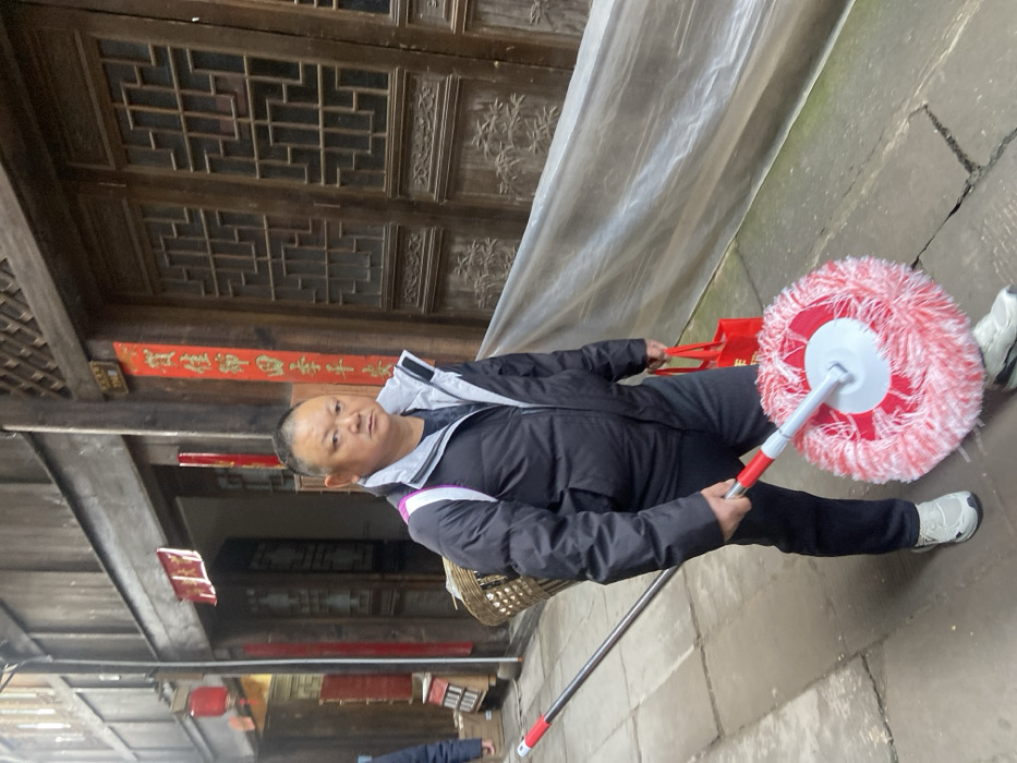 A man holds a mop with a red and white head. He wears a black jacket with a white collar, black pants, and white sneakers. He stands on a stone pathway next to a building with ornate wooden windows and a red banner. He carries a red plastic bag in his left hand, and a woven basket hangs off his left side. The image is rotated to the right.