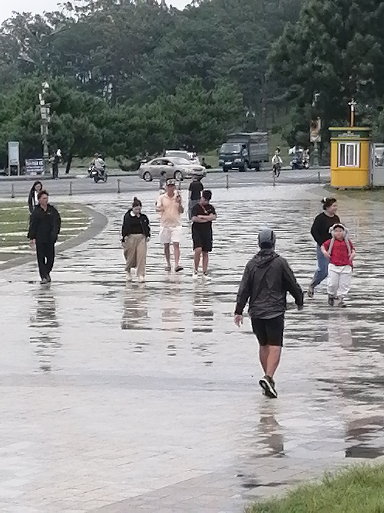 The image depicts a wet, paved outdoor area, likely a park or promenade, in Da Lat, Vietnam. Several individuals are walking across the reflective, rain-dampened surface. In the foreground, a person wearing a black hooded jacket and black shorts walks away from the camera. To the left, a person in a black jacket and black pants walks towards the camera. Further left, a person in a light-colored shirt and white shorts walks towards the left edge of the frame. In the mid-ground, a person in a black outfit with light-colored pants walks towards the right. Another person in a dark shirt and light-colored pants walks towards the right, accompanied by a child in a red shirt and white pants. In the background, several vehicles, including a truck and motorcycles, are visible on a road, and a yellow kiosk stands near the right edge. Lush green trees and foliage form a dense backdrop, indicating a natural setting within the city. The overall atmosphere suggests a recent rain shower, with the ground reflecting the overcast sky and surrounding elements.