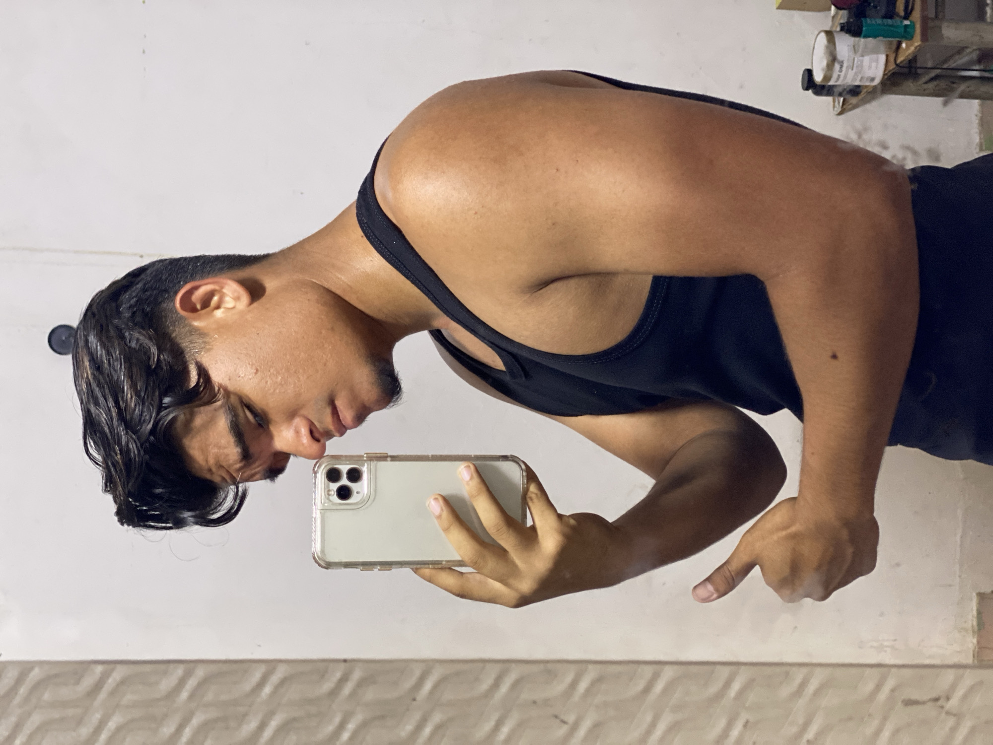 A man is taking a selfie in what appears to be an indoor setting. He is wearing a black tank top. The man is holding a smartphone, likely an iPhone, in his left hand while pointing with his right hand. Behind him is a white wall and a tiled section. The exact location in the city of Juan Griego, Venezuela, cannot be determined from the image.
