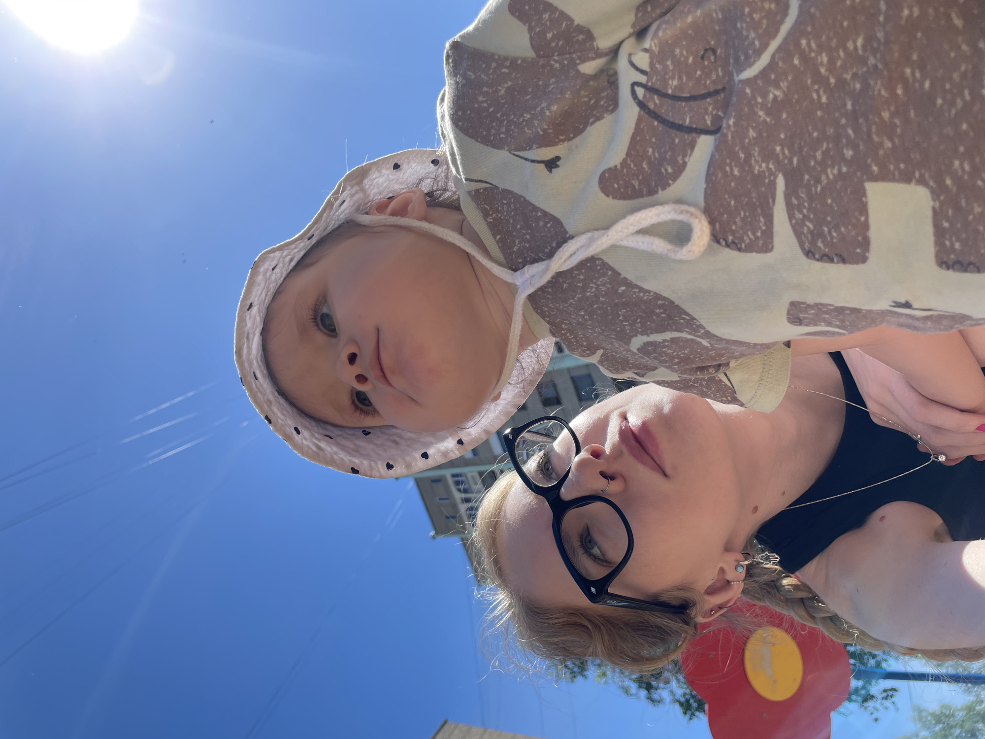 A low-angle photograph captures a woman wearing black-framed glasses, accompanied by a child wearing a patterned bucket hat. The blue sky fills the background. A partially visible building is in the background. The presence of playground equipment suggests the location is a park or recreational area in Izhevsk, Russia.