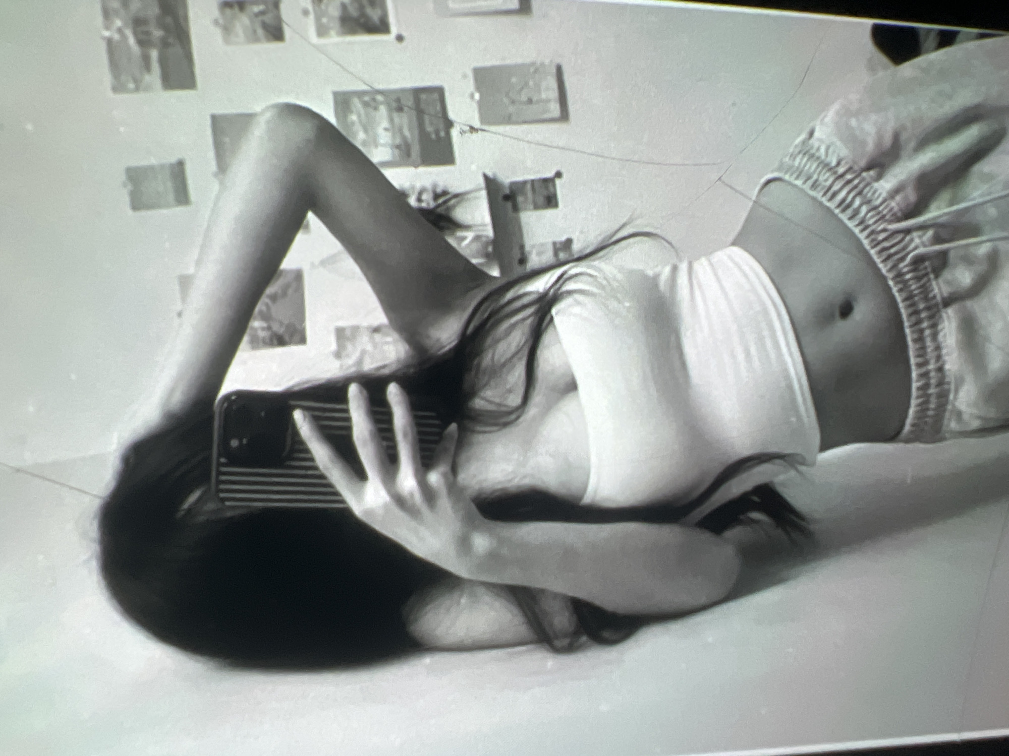 A black and white, slightly blurred, medium shot image shows a young woman taking a mirror selfie. She has long, dark hair that falls over her left shoulder and partially obscures her face. She is wearing a white, strapless tank top and light-colored sweatpants with a drawstring waist. Her right arm is raised, and her hand is playing with her hair. The background appears to be a bedroom, with a wall covered in small, framed pictures and a shelf. The overall lighting is soft, and the image has a grainy quality. The image does not contain any identifiable landmarks or features that would allow for determination of its location in Kara-Dıykan, Kyrgyzstan.