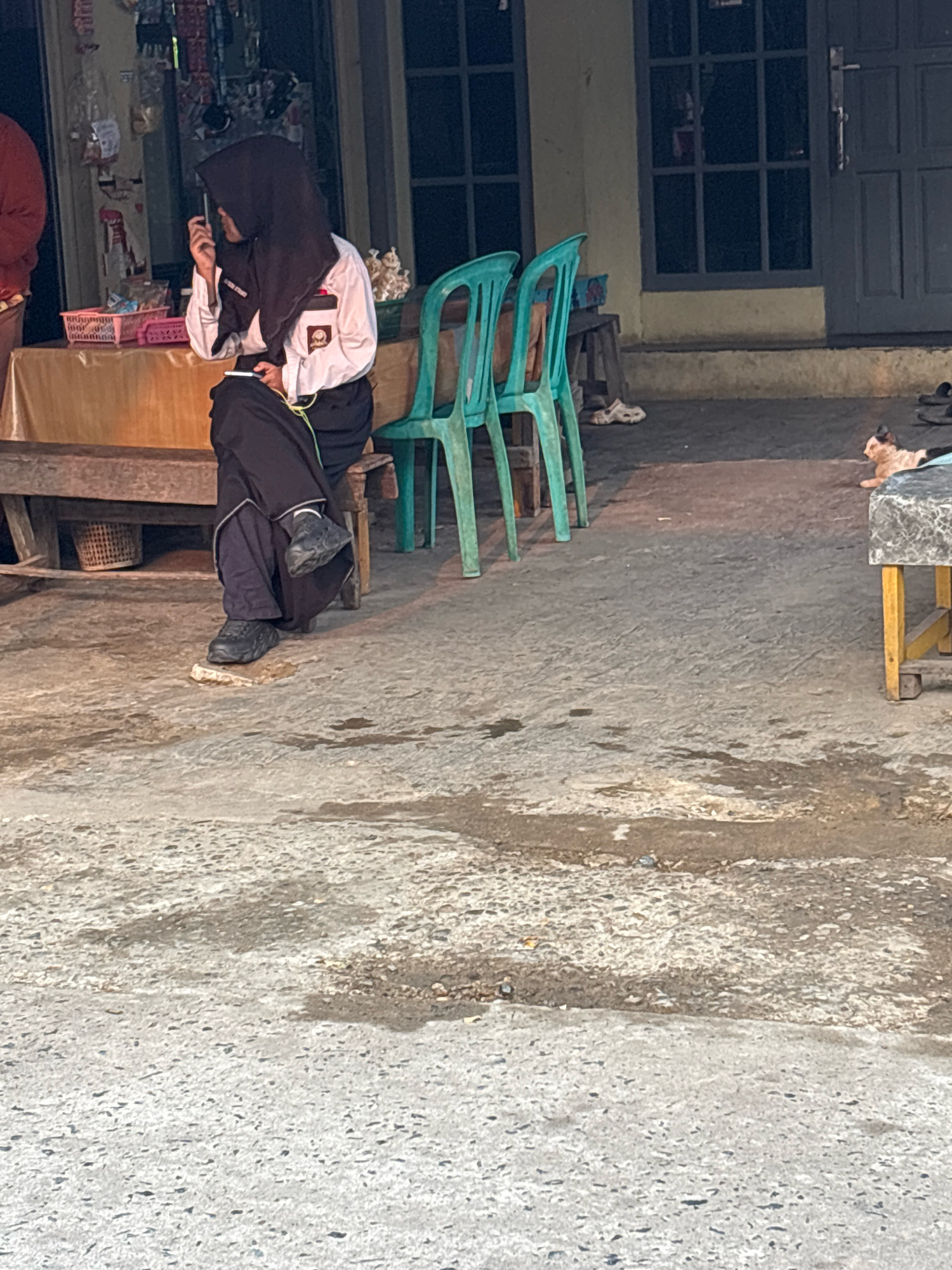 A young person wearing a black hijab and a white school uniform sits on a wooden bench in front of a building, holding a smartphone. The person is dressed in a long black skirt and dark shoes. Two teal plastic chairs are placed to the right of the bench. The ground is paved with rough concrete. In the background, the building has large windows and a dark door. A small, light-colored cat is lying on the ground to the right of the chairs, near a table with a patterned top. The scene appears to be outdoors, possibly on a street or in a public area in Banjarmasin, Indonesia, indicated by the architectural style and the presence of a school uniform.