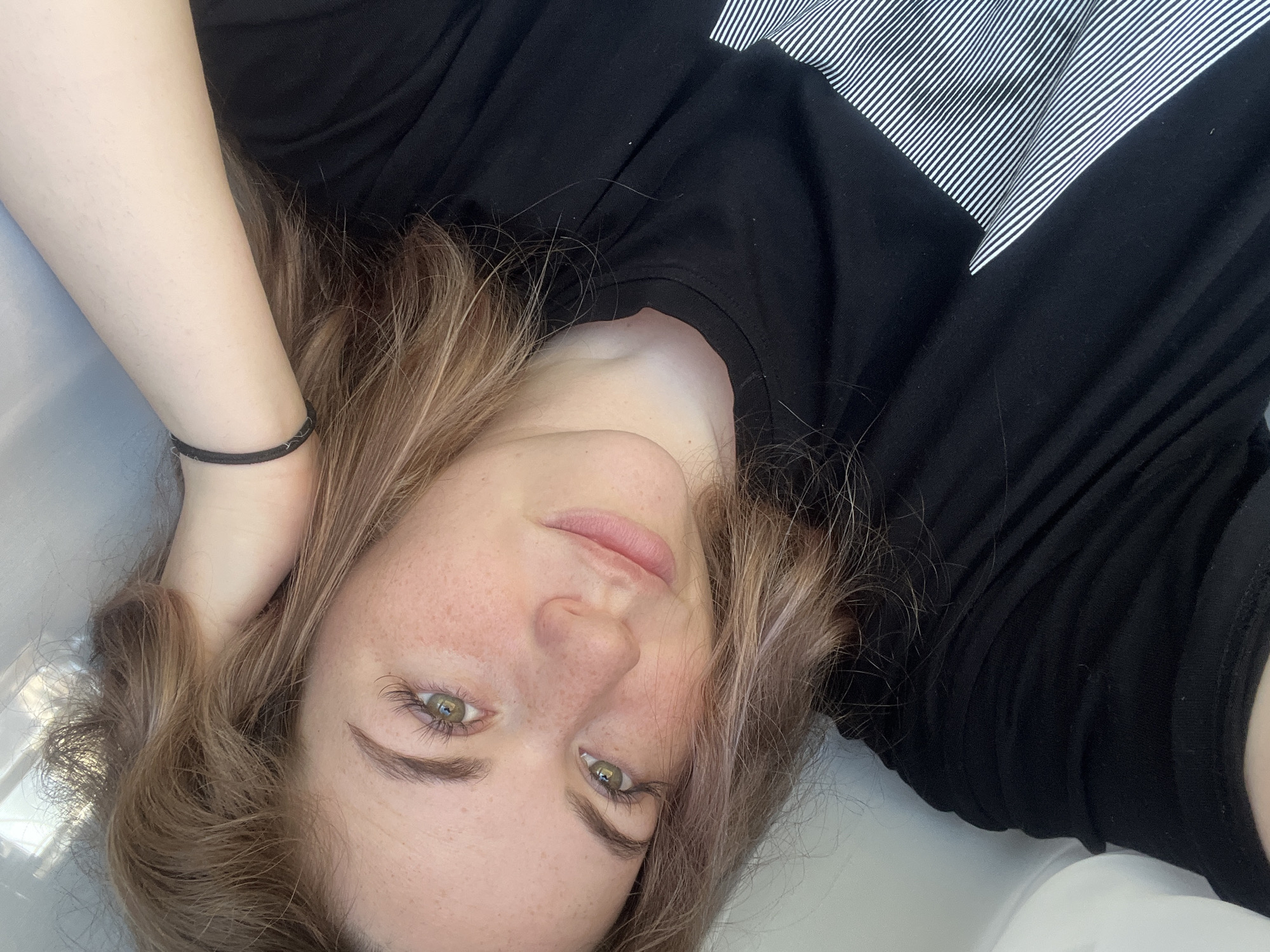 A young woman lies on her back in what appears to be a sink or bathtub, based on the white, curved surface beneath her. She has light brown hair with hints of pink, fair skin with freckles, and light green eyes. She's wearing a black long-sleeved shirt that partially covers another garment with black and white stripes. Her left arm is raised with her hand touching her hair. The location is not discernible from the image. There is no evidence in the image to indicate the city of Сосново, Russia.