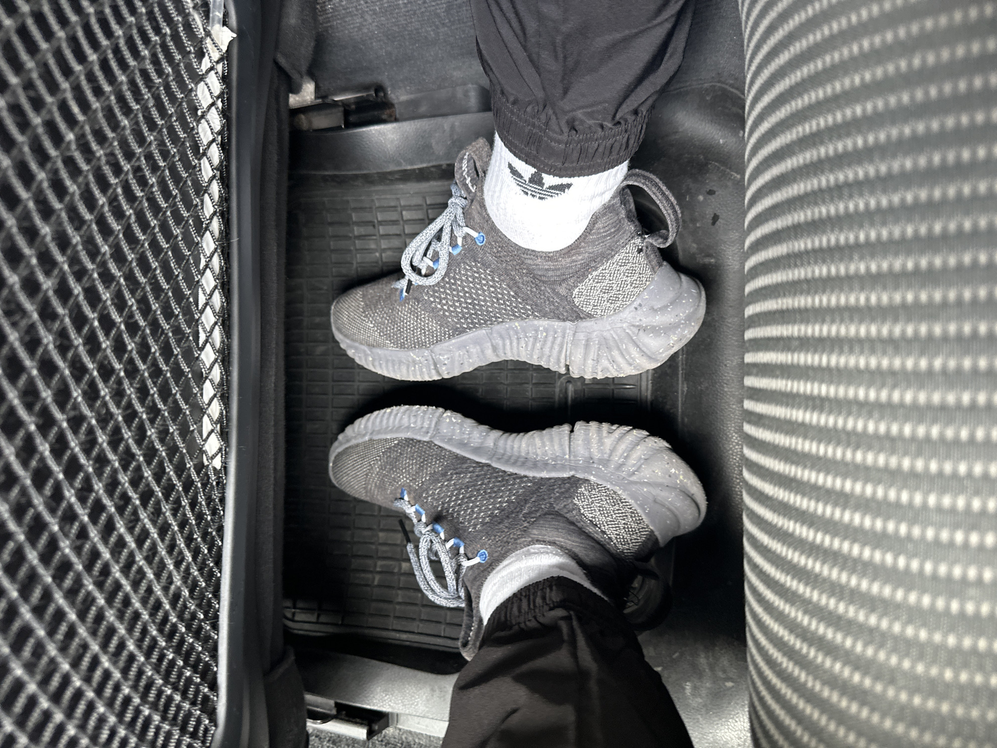 The image presents a downward, first-person view of two feet resting on a black rubber car floor mat. The feet are adorned with dark grey/black sneakers featuring light blue eyelets and speckled grey and white laces. The person wears white ankle socks, one of which prominently displays a black Adidas Trefoil logo. Black athletic-style pants, cinched at the ankles, cover the lower legs.
The scene is set inside a vehicle, likely a car or van, with the feet placed in the footwell. To the left, a black mesh storage net or pocket is visible, while to the right, a grey car seat fabric with a repetitive pattern of white dots fills the frame. The floor mat has a textured surface with raised patterns, typical for collecting debris. No specific activity is depicted beyond being present in the vehicle. The lighting suggests an interior environment. No emotions are discernible. The only visible text is the Adidas logo on the sock. The location is Zurzach, Switzerland.