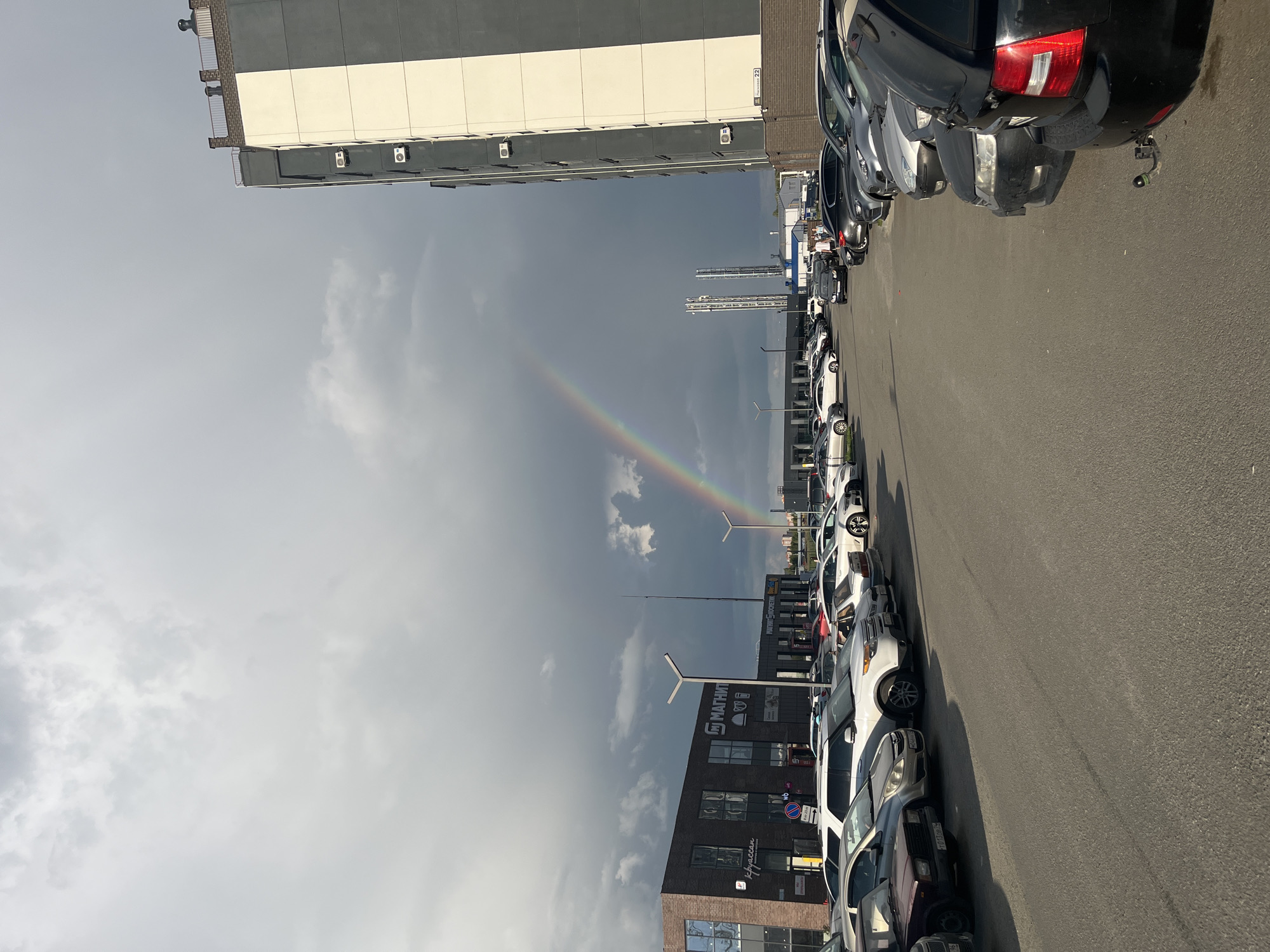 A rainbow arches across a dark, cloudy sky over a parking lot in front of a commercial building and a residential building. The parking lot is filled with parked cars. The commercial building has brick facade and multiple windows with signs. The residential building is tall and modern with a light-colored facade and balconies. The weather appears to be a mix of sun and rain, creating the rainbow.
