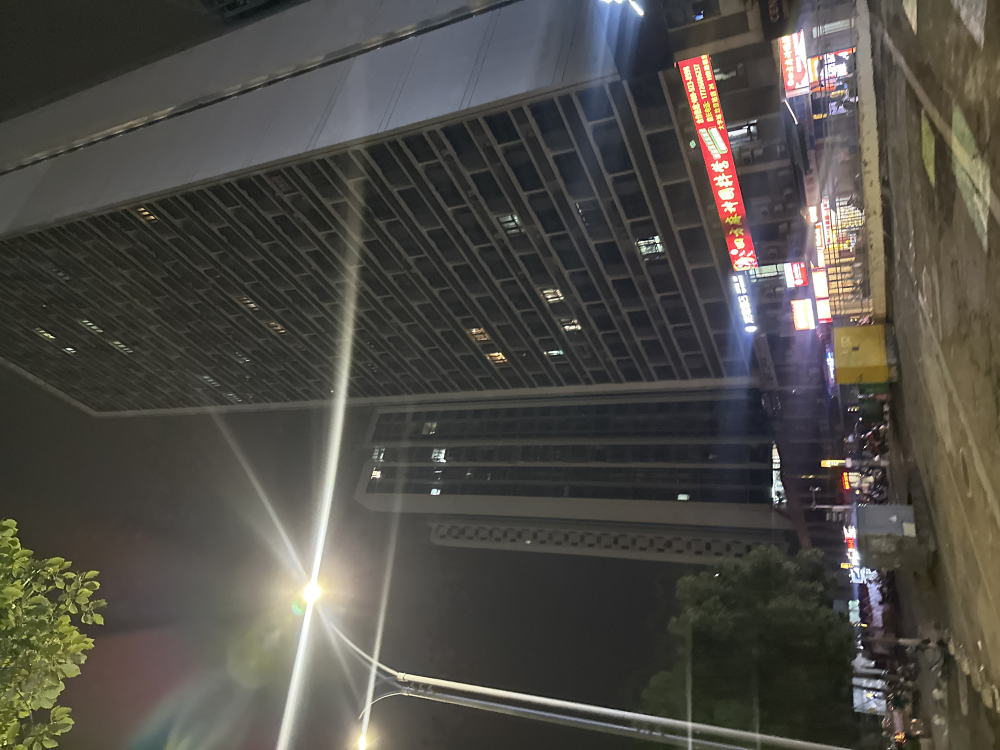 This is a nighttime street-level photograph of an urban environment in the Chongqing High-tech Zone, China. The composition is dominated by two tall, modern high-rise buildings. The building on the right is a dark, brick-patterned facade skyscraper, with numerous lit windows visible, indicating occupancy. The building on the left is a lighter-colored, more slender skyscraper, also with lit windows. In the foreground, a street is visible, with what appears to be wet pavement reflecting the city lights. There are several parked motorcycles and what might be a yellow utility box or small building in the lower part of the frame. A street lamp with bright lights and some lens flare is prominent on the left side of the image, casting light across the scene. Trees with green foliage are also visible in the lower left quadrant, their leaves illuminated by the artificial light. Commercial signs with Chinese characters are affixed to the lower levels of the buildings, some of which are brightly lit with red and white lettering, suggesting retail or service establishments. The overall atmosphere is that of a busy, illuminated city at night.