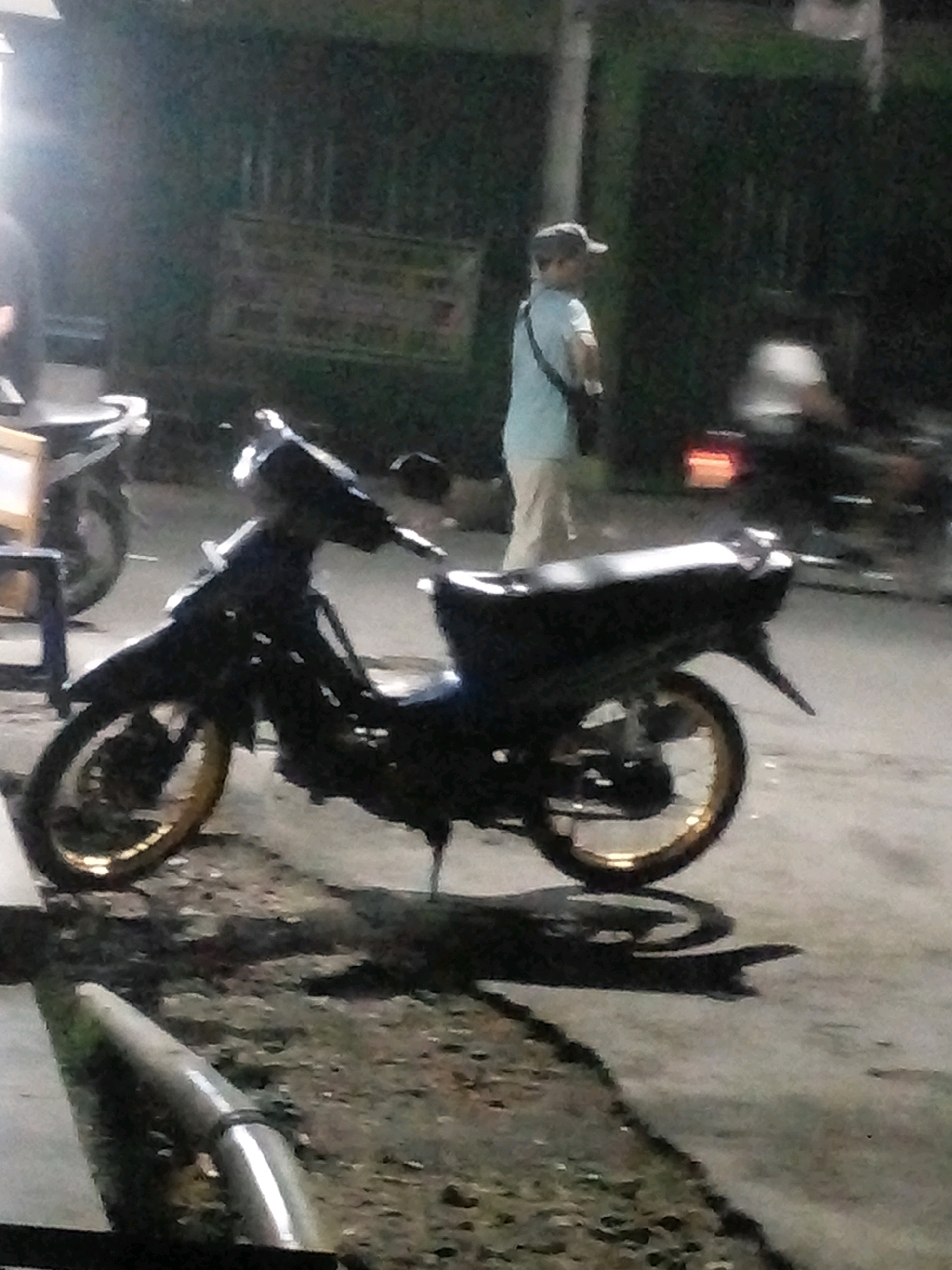 The image captures a nighttime scene in Limpung, Indonesia, featuring a parked motorcycle in the foreground. The motorcycle is dark in color with gold rims on both wheels. To the left of the motorcycle, a portion of a wooden structure is visible. In the background, a person wearing a light-colored t-shirt, beige pants, and a baseball cap is standing, with a bag slung across their body. Behind the person, another motorcycle is partially visible, with its taillight illuminated. The environment appears to be outdoors, possibly a street or roadside, with a dark, textured surface beneath the vehicles. The lighting suggests it is either dusk or nighttime, with some artificial light sources creating reflections and shadows.