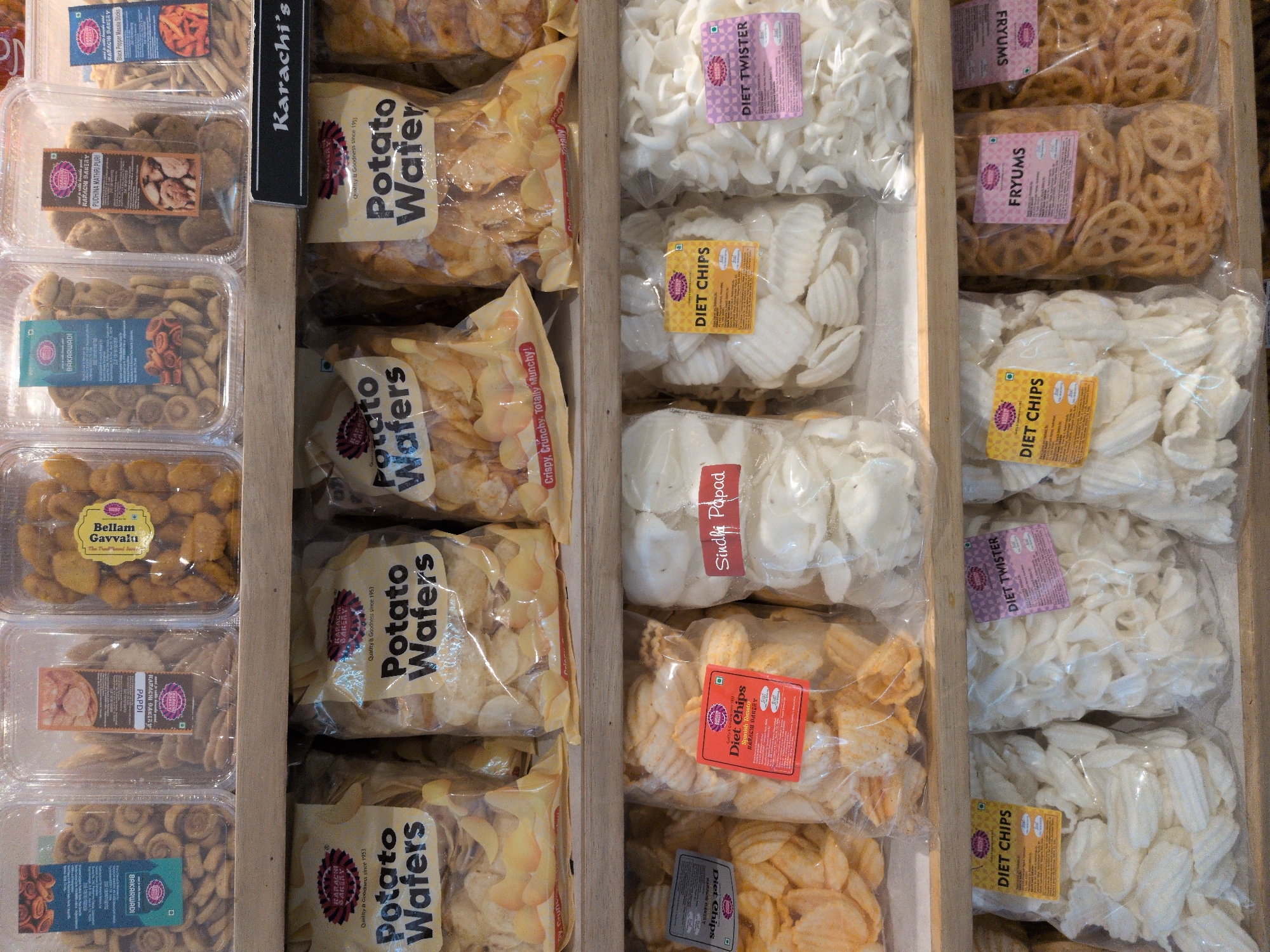 The image displays a retail store shelf unit featuring four tiers of packaged food products, predominantly from "Karachi Bakery," a brand established in 1953. Given the specific request for location, this scene is situated within a Karachi Bakery outlet or a retail section selling their products in Hyderabad, India.
The top shelf contains clear, rectangular plastic containers. From left to right, visible products include: "BAKARWADI," small, brown, spiraled snacks with a green vegetarian symbol; "PAPDI," flat, light brown, disc-shaped snacks; "Bellam Gavvalu," irregularly shaped, golden-yellow items; "PUDHINA MATURI PURI," dark brown, textured snacks; and partially visible "BLACK PEPPER MASALA STICKS," thin, elongated sticks. All these products carry the "Karachi Bakery" branding and typically a green vegetarian symbol.
The second shelf features a black sign with white lettering displaying "Karachi's" on the right side. Below this sign, three clear plastic bags of "Potato Wafers" are arranged side-by-side. Each bag contains light yellow, crinkle-cut potato chips and is labeled with "Karachi Bakery," "Quality & Goodness since 1953," and phrases like "Crispy, Crunchy, Totally Munchy!".
The third shelf holds various bagged snacks. From left to right: a partially obscured bag of wavy-cut chips; a bag of orange-yellow, crinkle-cut "Diet Chips" from Karachi Bakery, identified by a red label; a bag of white, irregular, shell-shaped items labeled "Sindhi Papad" directly on the bag; a bag of white, crescent-shaped "Diet Chips" from Karachi Bakery, with a yellow label; and a bag of white, twisted, spiral-shaped "DIET TWISTER" from Karachi Bakery, identified by a purple label. All Karachi Bakery branded products on this shelf feature a green vegetarian symbol.
The fourth and bottom-most visible shelf continues the display of bagged products. From left to right: a bag of white, irregular, shell-shaped "Diet Chips" from Karachi Bakery, with a yellow label and green vegetarian symbol (identical to one on the third shelf); a bag of brown, circular, lattice-patterned snacks labeled "FRYUMS" from Karachi Bakery, with a purple label and green vegetarian symbol; and another identical bag of "FRYUMS" to its right.
The shelves are constructed of light-colored wood. No individuals are present in the frame. The lighting is uniform, suggesting an indoor commercial environment.