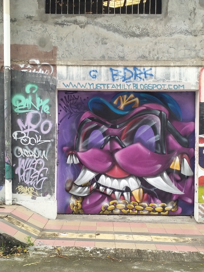 This media file presents a vibrant and detailed graffiti artwork on a metal shutter within an urban setting in Karanganyar, Indonesia. The central piece is a large, stylized depiction of an aggressive-looking character or monster head, dominated by shades of purple and magenta. It features prominent, intense dark eyes, an open maw revealing numerous sharp white fangs, some tipped with gold, and a heavy golden chain encircling its lower jaw. A blue baseball cap with a distinct yellow emblem (possibly a stylized 'NY') sits atop its head.
Above the main artwork, blue graffiti reads "G BDRK" and "WWW.YUSTFAMILY.BLOGSPOT.COM". To the left, a concrete wall is heavily tagged with multiple layers of graffiti in various colors, including light green, white, and grey. Visible words and tags include "MOO", "ROCK", "WILD RULER", and "BANOR", amidst other less legible script.
The scene is an outdoor public space, likely a street or alley. The walls are made of weathered concrete, and an upper window is fitted with metal bars. A tiled sidewalk with yellow and pink stripes occupies the foreground, suggesting a pedestrian area. The image is captured during daylight, indicating clear weather. The overall impression is one of a dynamic urban landscape adorned with street art.