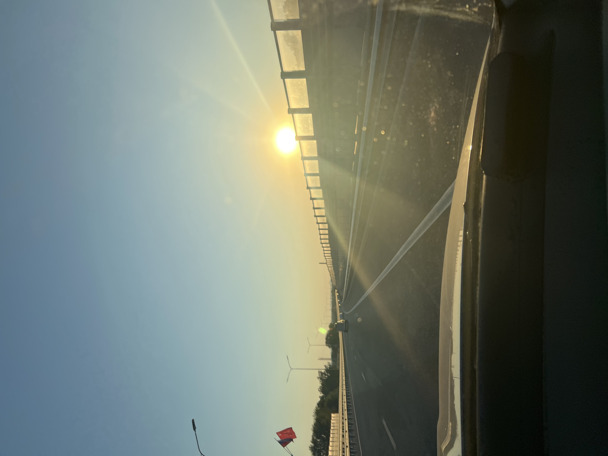 The image depicts a view from a car driving on a highway at sunrise. The sun is low on the horizon, casting a warm, golden light and creating lens flare effects. The highway stretches ahead, with lanes marked by white lines. On the right side of the highway, there is a barrier with transparent panels, beyond which a landscape is visible, partially obscured by the sun's glare. On the left side of the highway, there are streetlights and some trees. Two flags, one red and one dark-colored, are visible on the left side, mounted on a pole. The dashboard of the car is visible in the foreground, reflecting the light. The sky is clear and blue, transitioning to a lighter hue near the horizon. There are no identifiable people or specific buildings that would pinpoint the location to Noven'kii, Russia.