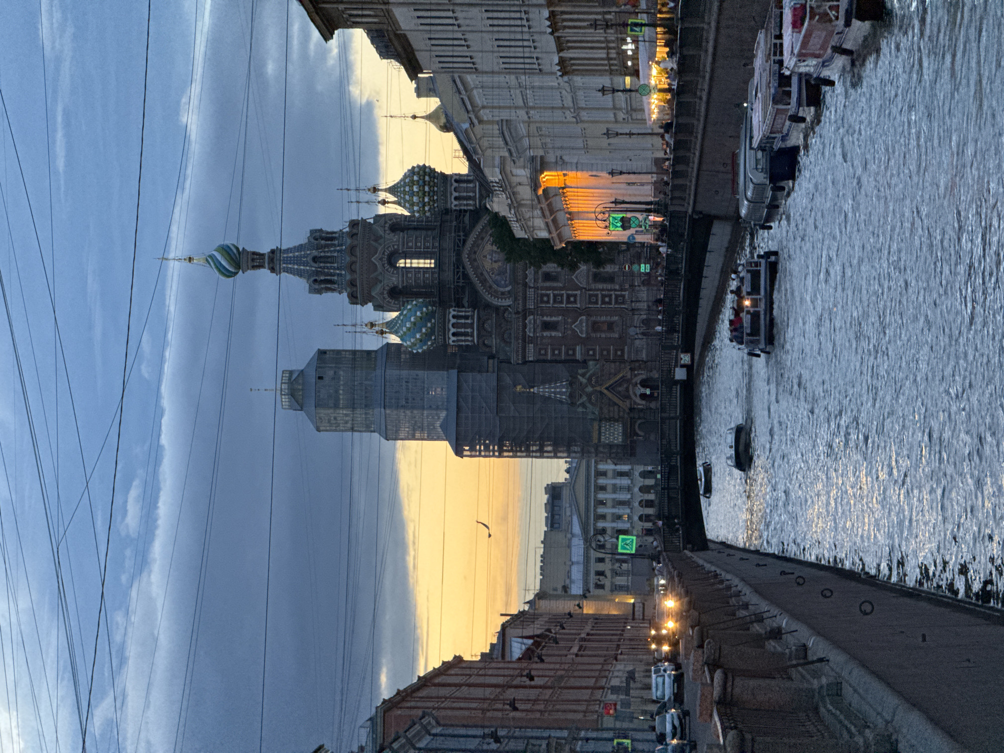This image captures a scenic view of the Griboyedov Canal in Saint Petersburg, Russia, at dusk. The iconic Church of the Savior on Spilled Blood is prominently featured with its distinctive onion domes, bathed in the warm glow of the setting sun. Several tour boats are navigating the canal, carrying passengers who are enjoying the evening views. The canal banks are lined with historic buildings, their windows reflecting the fading light. Streetlights and illuminated storefronts add to the lively atmosphere. The sky above is a dramatic blend of twilight blues and oranges, with a few wisps of clouds. Overhead power lines crisscross the sky, a common sight in urban landscapes. The overall scene conveys a sense of tranquility and urban charm.