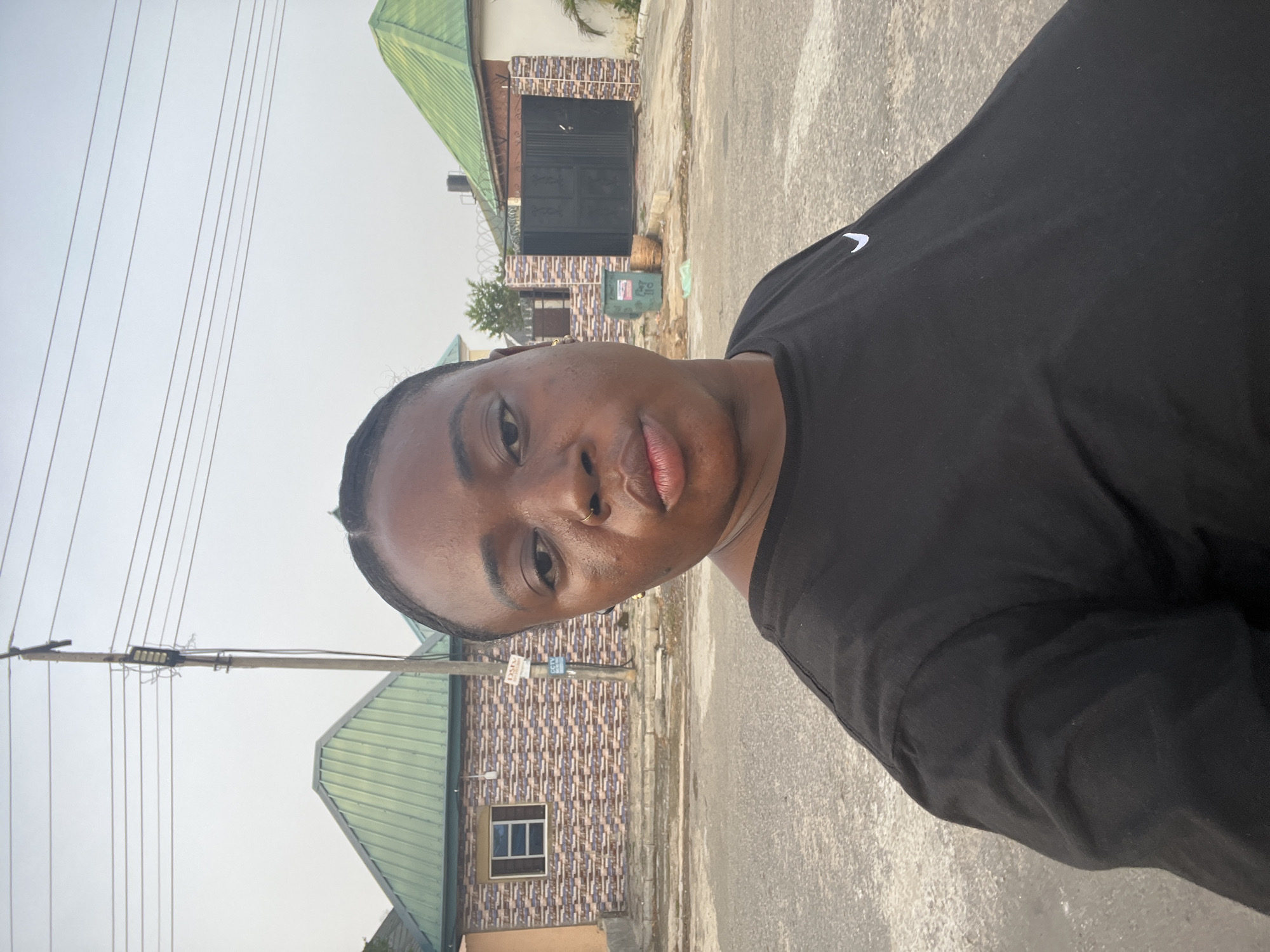 A medium shot shows a person in a black shirt, standing outside. The person's face is well-lit, and they are looking directly at the camera. The background includes a street, houses with green roofs, and utility poles with wires. The image could be in Abuja, Nigeria, based on the architectural style of the houses, which is typical of the region.