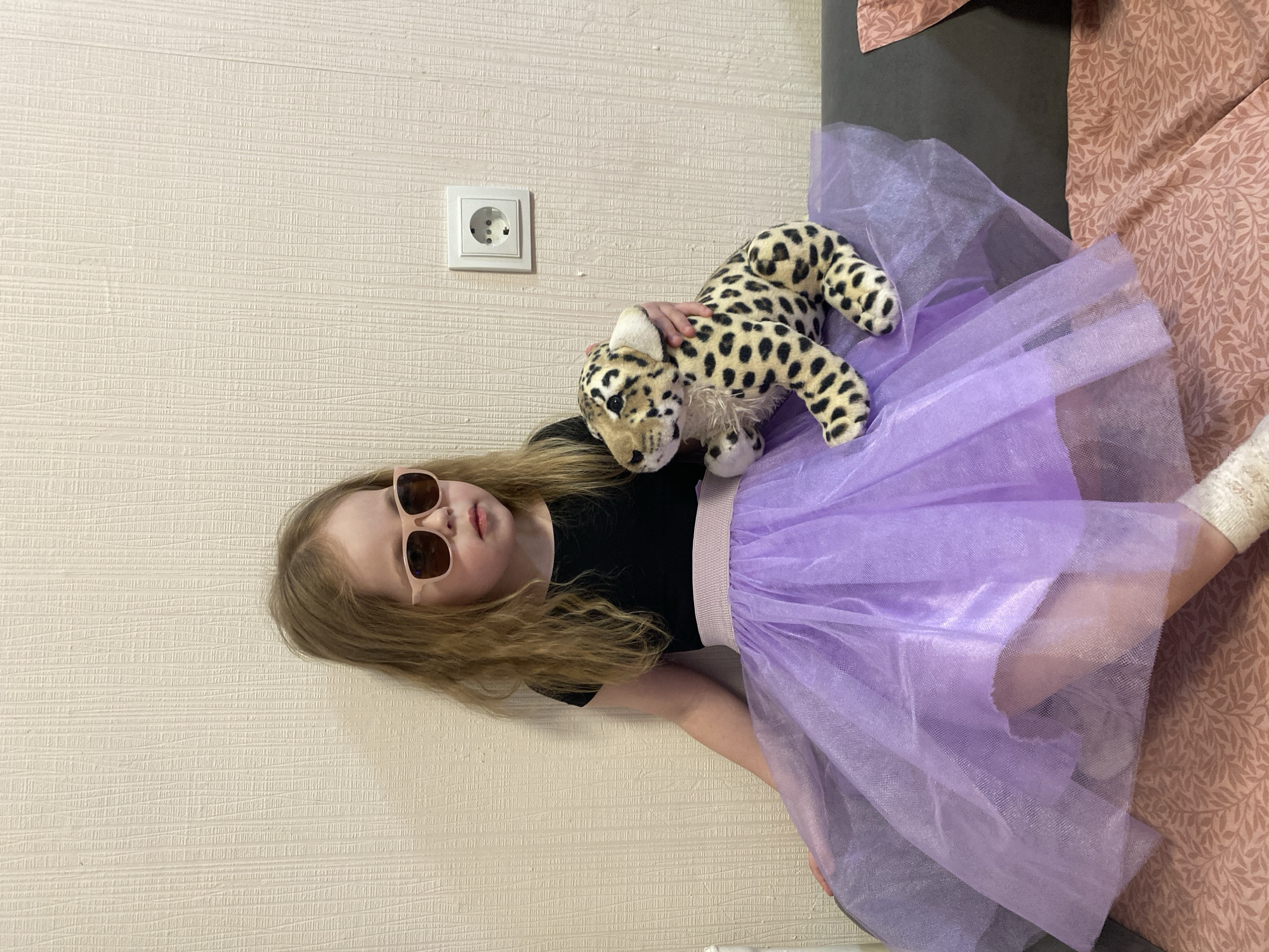 A young girl is pictured indoors, reclining or lying against a textured, light-colored wall, possibly in a home in Izhevsk, Russia. Her head is positioned towards the upper left, and her body extends diagonally across the frame.
The girl has light brown hair and is wearing a black t-shirt and a fluffy, sheer purple tulle skirt. Her eyes are concealed by light-framed, dark-lensed sunglasses. She is holding a spotted stuffed animal, resembling a leopard or cheetah, which rests on her midsection. Her right hand is visible on the stuffed animal's head.
On the wall behind her head, a white electrical outlet with two circular sockets and a grounding pinhole is visible, consistent with a European-style (Type F/Schuko) socket found in Russia. To her right, part of a bed or sofa covered with a pinkish-brown fabric featuring a repeating leaf pattern is visible. The scene is illuminated by indoor lighting, suggesting daytime or artificial light. The girl appears to be posing in a relaxed, playful manner. No text is visible in the image.