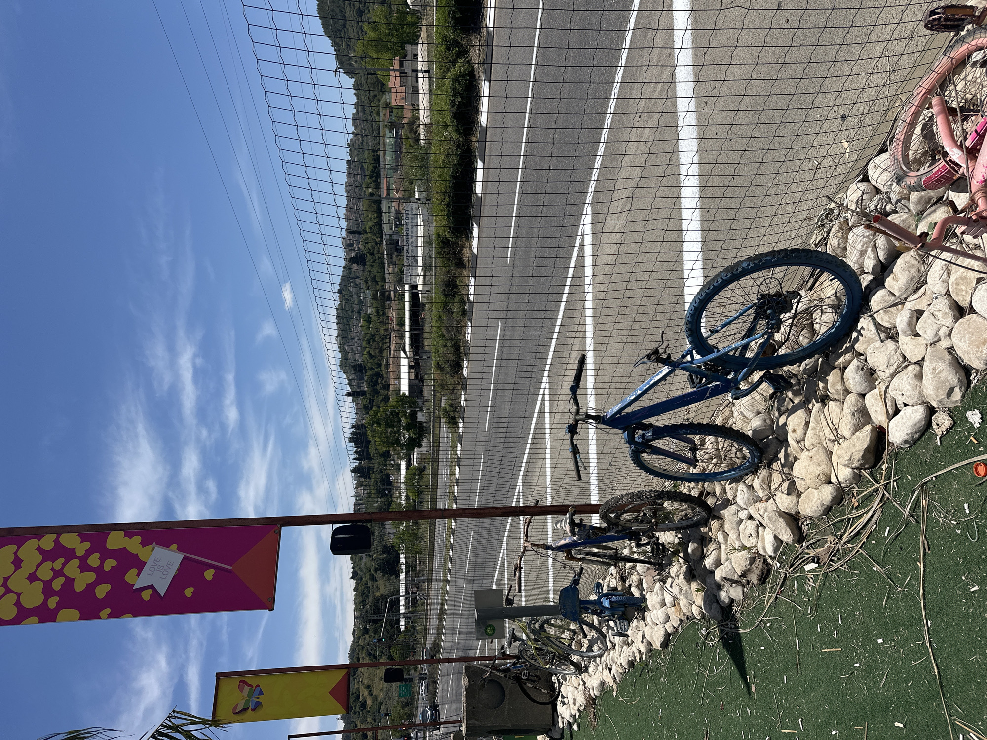 The image shows a roadside scene in Maalot-Tarshiha, Israel. A chain-link fence separates a line of parked or abandoned bicycles from a roadway. The roadway features white lines marking lanes. Beyond the road, a vegetated hillside rises. The foreground also contains banners with decorative and symbolic designs. The bicycles are of various types and conditions, some resting on a bed of rocks and artificial turf. The weather appears clear and sunny.