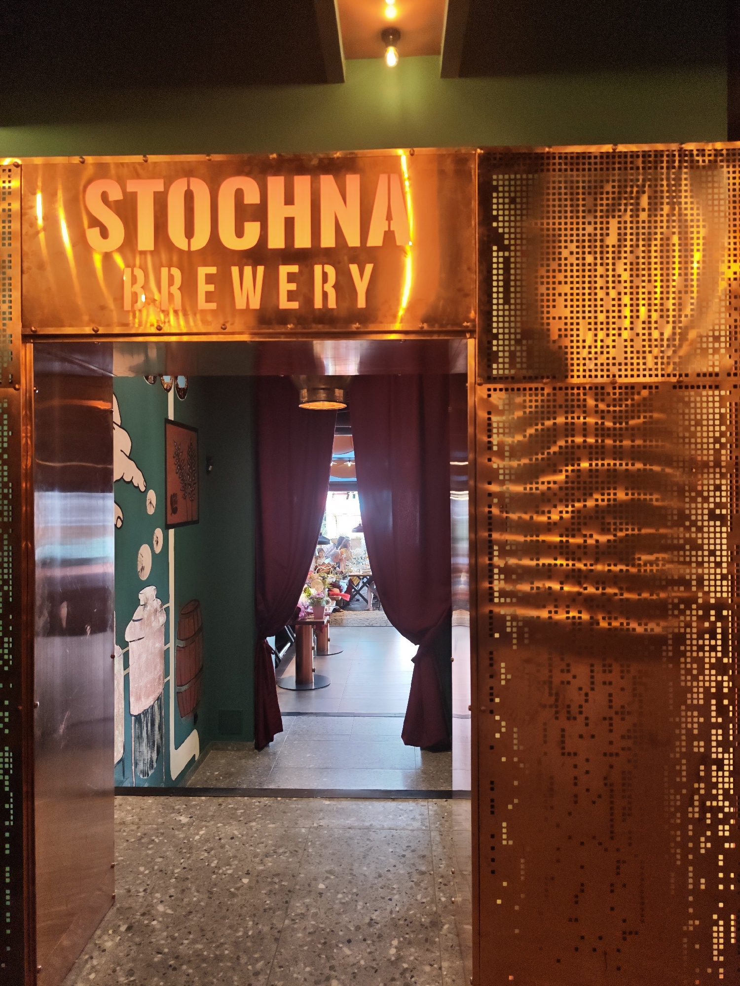 The image presents the entrance to Stochman Brewery in Sofia, Bulgaria. A prominent overhead sign, constructed from a luminous coppery-bronze metal, displays "STOCHNA BREWERY" in glowing orange, stencil-like lettering. The entrance portal itself is framed by the same metallic material; the left panel is solid, while the right panel is perforated with a repeating square grid pattern, reflecting and emitting warm orange light. Above the sign, a dark ceiling transitions into a green wall. A single exposed incandescent bulb fixture is visible on the ceiling.
Through the entrance, a short hallway extends into the establishment. The immediate left wall inside the entrance is painted a teal-green and adorned with stylized white murals depicting a large hand, circular motifs, a wooden barrel, and a substantial container or bottle. A dark-framed piece of art hangs on this wall. Heavy, dark red curtains are drawn back, framing the view into a brightly lit interior space. The floor throughout the visible area is a speckled terrazzo. Beyond the curtains, partial views of a dining area show tables with dark tops and light bases. Several indistinct human figures are seated at these tables. A small bouquet of flowers rests on a table in the foreground.