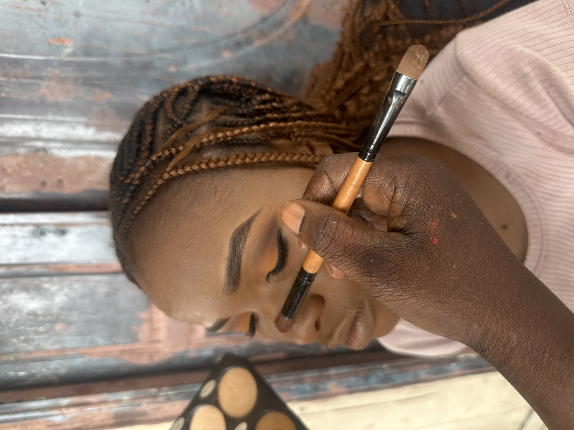 A medium shot shows a Black person's face from the nose up, with their eyes closed, as a makeup artist applies eye shadow with a brush. The makeup artist's hand, with visible wrinkles and some smudged makeup, holds the brush and is positioned near the subject's nose. A makeup palette with various foundation shades is visible in the upper left corner. The subject has braided hair with brown highlights, and is wearing a light pink ribbed t-shirt. The background is a textured, metallic surface, possibly a corrugated metal door, with patches of rust and peeling paint. The scene appears to be set in an indoor or covered area, possibly a salon or a home, in Badagry, Nigeria, indicated by the style of the makeup and the texture of the background which is common in older structures in the region.