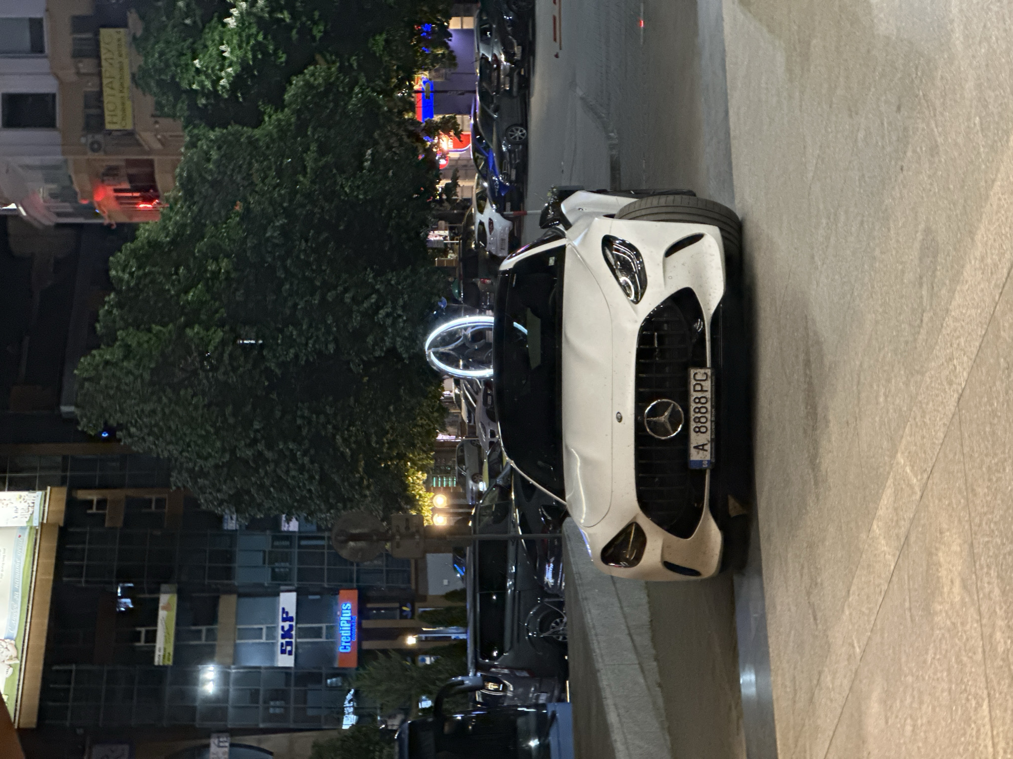 The image shows a white Mercedes-Benz AMG GT parked on a street in Plovdiv, Bulgaria. The car has a Bulgarian license plate that reads "A 8888 PC". The street is lined with buildings, some of which have signs in Bulgarian, including signs for "SKF" and "CrediPlus." A sign for "НОТАРИУС Стоянка Кисьова" is visible as well. The scene is at night, and streetlights illuminate the area. There are other cars parked along the street. A circular light installation is visible in the background.
