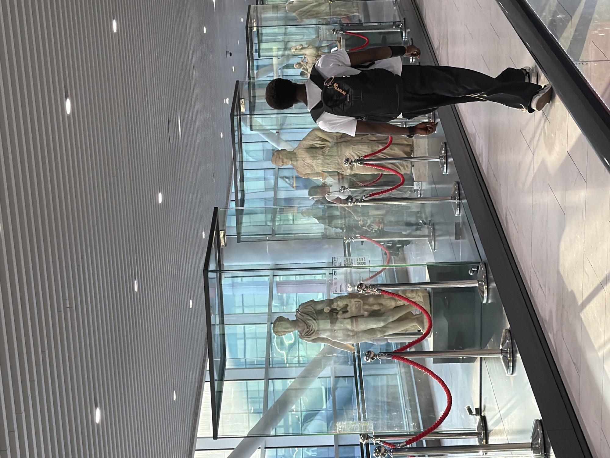 The image depicts a contemporary indoor exhibition space, likely within a modern building or airport terminal in Fiumicino, Italy. In the foreground, a Black man with short, dark hair walks away from the viewer, wearing a white t-shirt, black pants, and a black backpack. He is walking on a moving walkway.
The background features several glass display cases containing marble statues. One statue in the foreground, partially visible, is a classical male figure. Further back, other statues are visible, including what appears to be a female figure and a horse statue. Red velvet ropes with chrome stanchions are used to cordon off the exhibits. The environment is well-lit with overhead lighting, and large windows with blue-tinted panes are visible in the background, reflecting sunlight and the surrounding architecture, suggesting a connection to the outdoors. The ceiling is a modern, slatted design. The overall impression is one of a cultural display in a public, modern setting.