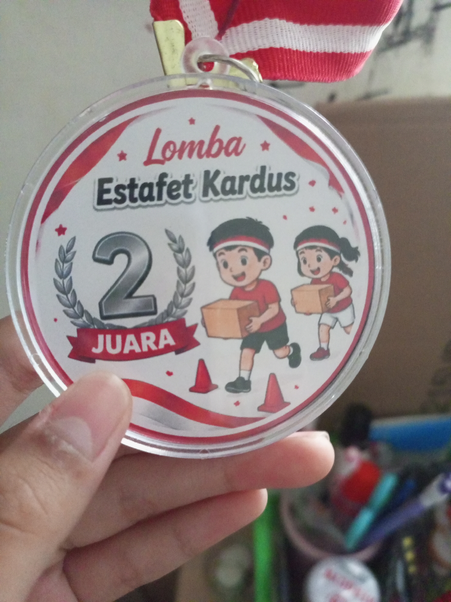 A circular medal with a red and white ribbon hangs from a thin gold loop. The medal features an illustration of two cartoon children, one boy and one girl, wearing red shirts and headbands with black and white stripes. They are both carrying brown boxes. The boy is on the left and the girl is on the right, and they appear to be running. In front of them are two orange traffic cones. The medal has text that reads "Lomba Estafet Kardus" in English "Cardboard Relay Race." To the left of the children, a large silver number "2" is surrounded by a laurel wreath, and beneath it, a red banner reads "JUARA," which translates to "Champion" in English. Red stars are scattered across the white background of the medal. The medal is being held up by a person's hand, with fingers and thumb visible. The background is blurred and appears to be indoors, with some shelves and containers of colorful items like pens visible. The location is not specified.