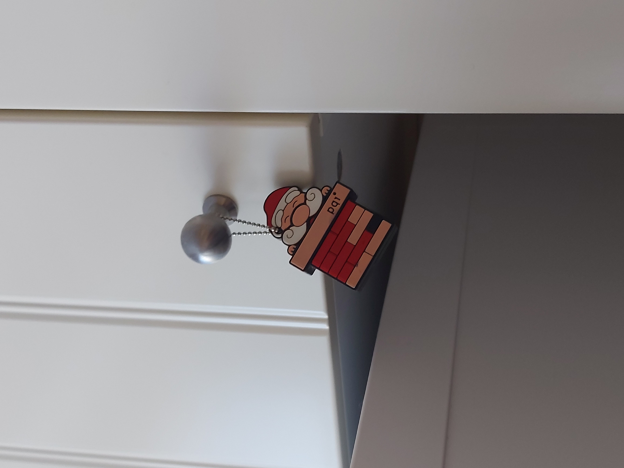 A close-up photograph depicts a Santa Claus keychain hanging from a silver knob on a white drawer. The keychain features a cartoon Santa figure partially inside a chimney, with "pqi" printed on the side of the chimney. The drawer has horizontal grooves running along its front. The backdrop consists of a gray surface. The location is not identifiable as Saint Petersburg, Russia, based on the visual information in the image.