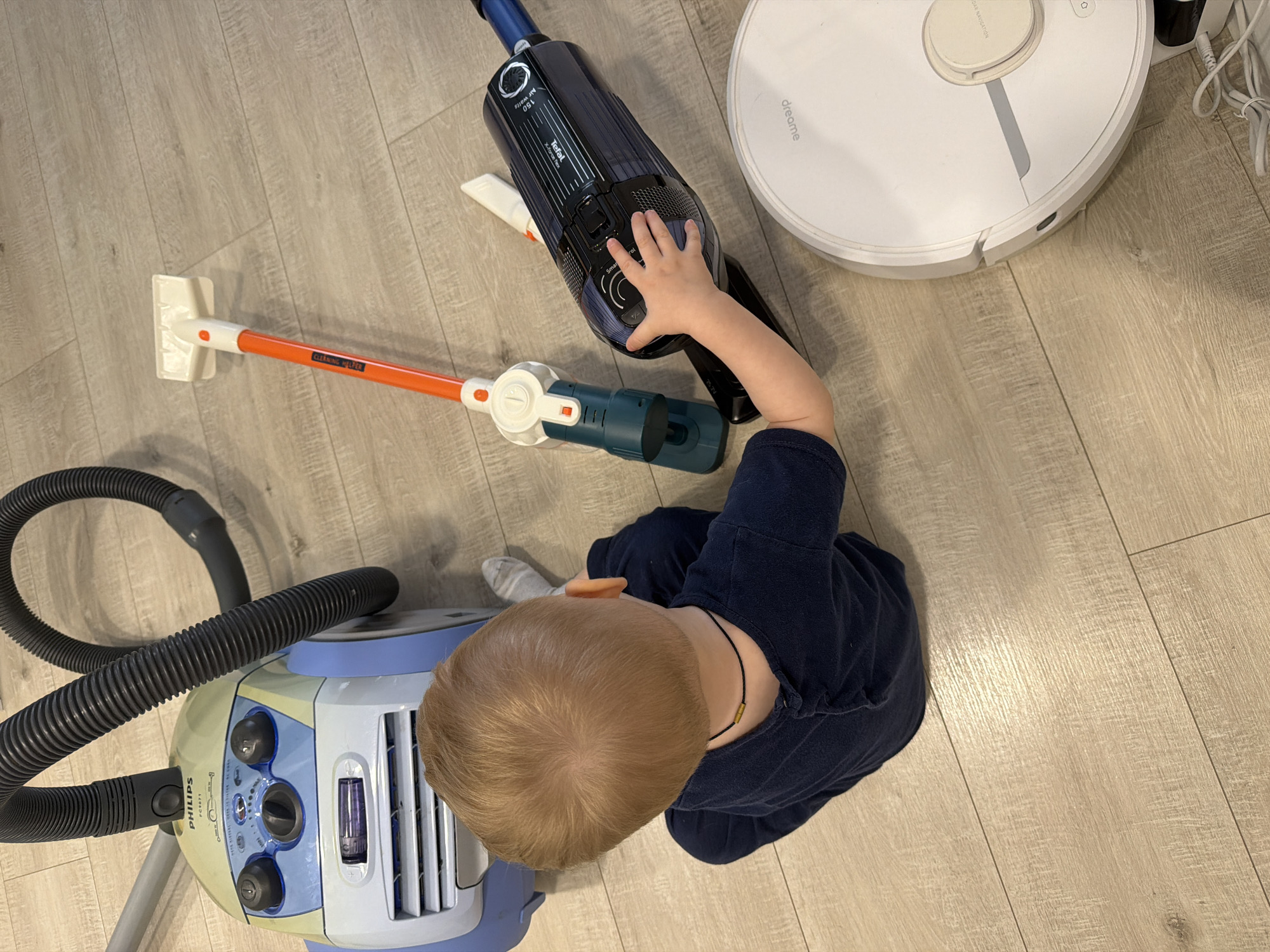 A young child with blonde hair, seen from an overhead perspective, interacts with several cleaning appliances on a light wooden or laminate floor indoors, in what is identified as Votkinsk, Russia. The child, wearing a dark blue top and grey socks, is reaching out with their right hand to touch a dark blue stick vacuum cleaner.
The scene features a collection of cleaning devices. Prominently displayed are:
- A dark blue stick vacuum cleaner, identified by "Tefal X-Force Flex," with "Air watts 150" visible on its body. The child's hand is on a control panel area that partially shows "Sm" and "+/-".
- A white circular robot vacuum cleaner, labeled "dreame," positioned to the upper right. Text "DAR NAVIGATION" is partially visible on its surface.
- A light blue and grey canister vacuum cleaner with a flexible black hose, located to the lower left. It is clearly marked "PHILIPS FC6071".
- An orange and white toy or child-sized cleaning stick, with "CLEANING HELPER" printed on its handle, lies on the floor near the child.
The overall impression is of a child engaging in playful exploration or learning about household cleaning tools. The setting appears to be a bright, casual indoor environment.