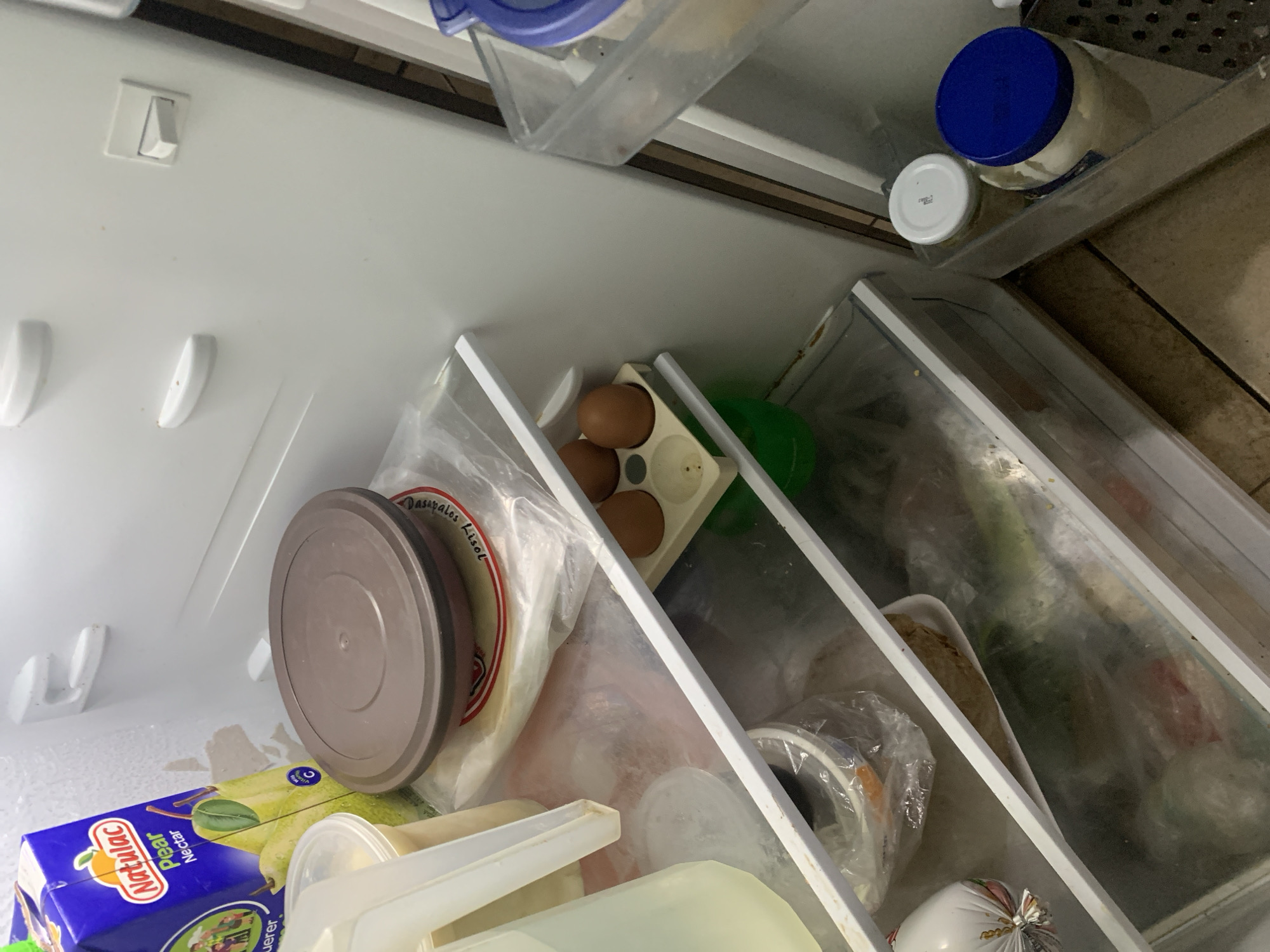 A high-angle, tilted photograph provides a view into the interior of an open refrigerator. The white walls of the appliance are brightly lit, revealing various food items and containers.
On the main shelf, a blue and yellow carton labeled "Natulac Pear Nectar" is prominent. Nearby, several plastic containers are visible; one has a brown lid, while another is a clear plastic bag containing flat, round items, possibly flatbreads, with the partial text "Dasapalos Lisiol" (or similar) visible. An egg tray holds four brown eggs, and a green bowl or container is tucked behind them. On the far left interior door panel, multiple white plastic handles or supports are attached. A white light switch is visible on the upper left wall of the refrigerator compartment.
The top shelf on the right side contains a large jar with a blue lid, likely a condiment, and a smaller jar with a white lid. This shelf and a clear plastic container nearby show some signs of use or minor discoloration. Below this, a translucent crisper drawer holds several items, mostly obscured by plastic bags and the drawer's frosted surface. Within the drawer, a white foam tray with a wrapped food item and a white plastic bag tied at the top, possibly containing cheese or sausage, can be discerned.
No people are present, and no specific activity is taking place; the image is a static snapshot of a refrigerator's contents. The setting appears to be a home kitchen in Santa Ana, Venezuela, suggested by the typical household items. The time of day is not determinable from the artificial indoor lighting.