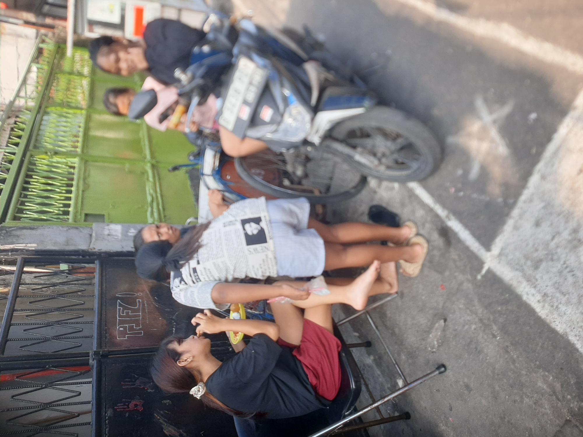 Four young women are gathered outdoors near a metal gate and a green fence. One woman, seated on a black folding chair, wears a black t-shirt and a maroon skirt. She is eating from a yellow plate. To her right, another woman, wearing a white shirt with text and light blue shorts, stands with her back to the camera, holding a clear plastic bag. Two other women are partially visible behind them, one wearing a pink top and the other a black long-sleeved shirt. A blue motorcycle is parked beside them, with its front wheel visible. The ground is paved, and there are markings on the concrete, including a white star shape. The overall setting appears to be a street in Central Jakarta, Indonesia, possibly near a residential area given the gate and fence.