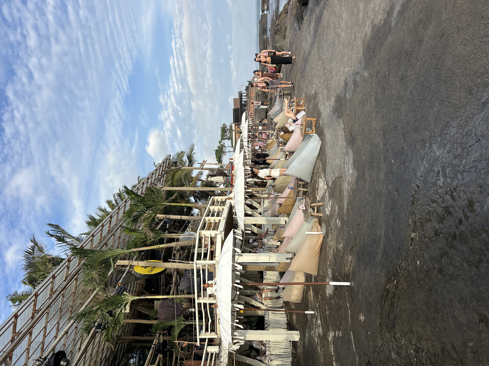 This image captures a vibrant beachside scene at La Brisa in North Kuta, Bali, Indonesia, on a clear day with scattered clouds. The foreground features a row of large, white parasols providing shade over a collection of multi-colored beanbag chairs, some occupied by people lounging or looking at their phones. Small wooden tables with low stools are interspersed among the seating.
Behind the beanbag seating area, a multi-story structure with a rustic, open-air design is visible. This building appears to be a restaurant or bar, with people seated on its upper levels. Palm trees are strategically placed, adding to the tropical ambiance. A prominent yellow circular sign with black lettering, possibly a logo or brand name, hangs from one of the support beams.
Further down the paved walkway, several individuals are seen walking along the rocky coastline. The sea is visible in the background, with waves gently lapping against the shore and rocks. The overall atmosphere is relaxed and suggests a popular tourist destination. The ground appears to be wet in some areas, possibly from recent tides or cleaning.