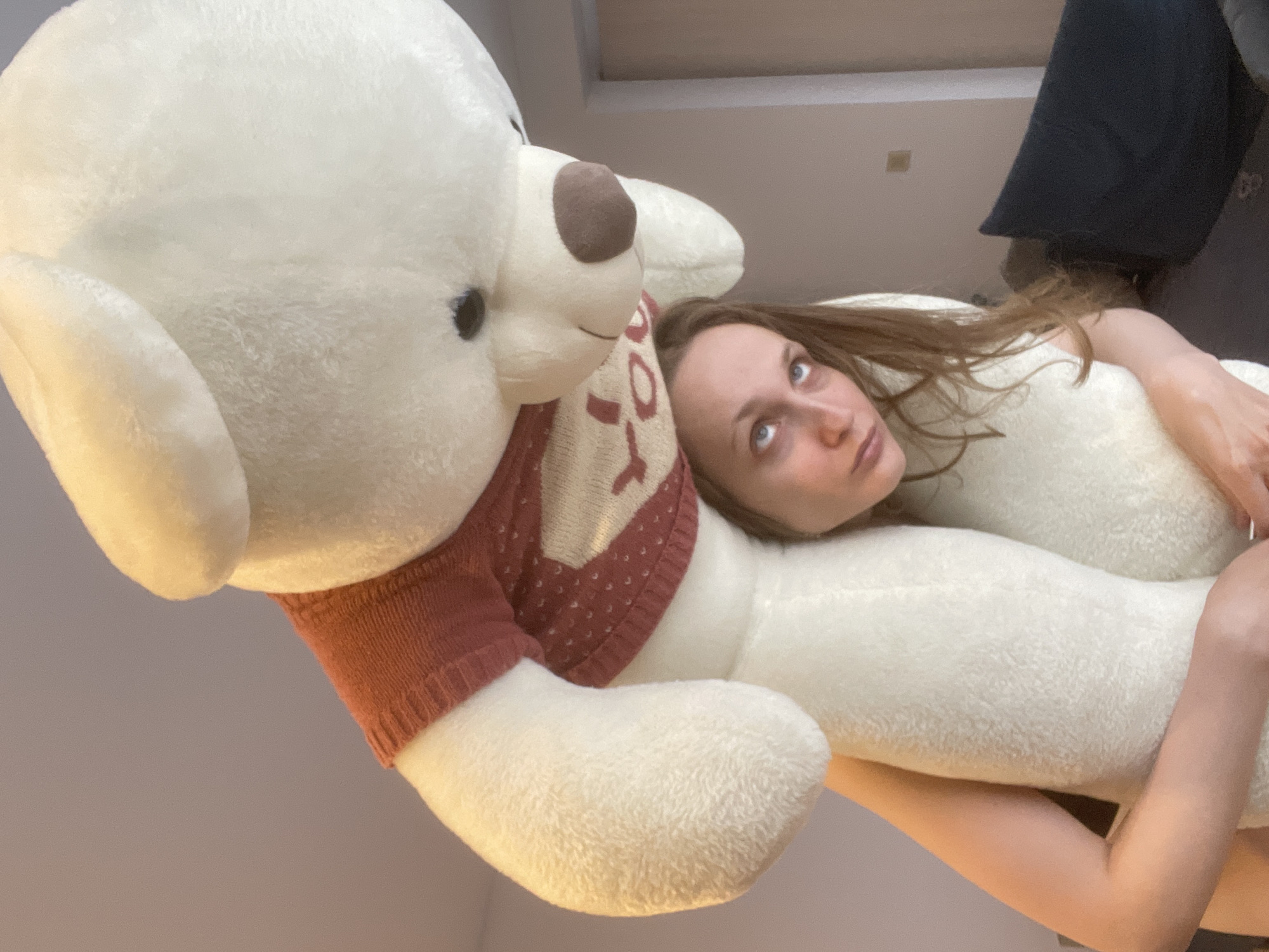 The image shows a young woman lying down and embracing a very large, white teddy bear in an indoor setting. The camera angle is tilted, making the woman and bear appear diagonally in the frame against a plain, light-colored background.
The primary subjects are the woman and the teddy bear. The woman has long, light brown hair, fair skin, and light-colored eyes, and she is looking upwards and slightly to the right with a neutral to slightly pensive expression. Her right arm is visible, wrapped around the plush toy, and her head rests on its body. The teddy bear is significantly large, with soft white fur, dark eyes, and a brown nose. It is wearing a knitted sweater in red and white with a patterned design on the front, which appears to be a stylized shape rather than discernible text.
The scene takes place indoors, likely in a private room such as a bedroom or living room, suggested by the plain wall/ceiling and what appears to be a dark piece of furniture or bedding in the upper right background. The lighting is soft and even, typical of indoor artificial light or diffuse daylight. The overall atmosphere conveys comfort, relaxation, and affection. The specific location context is Подшивалово, Russia. There is no visible text in the image.
