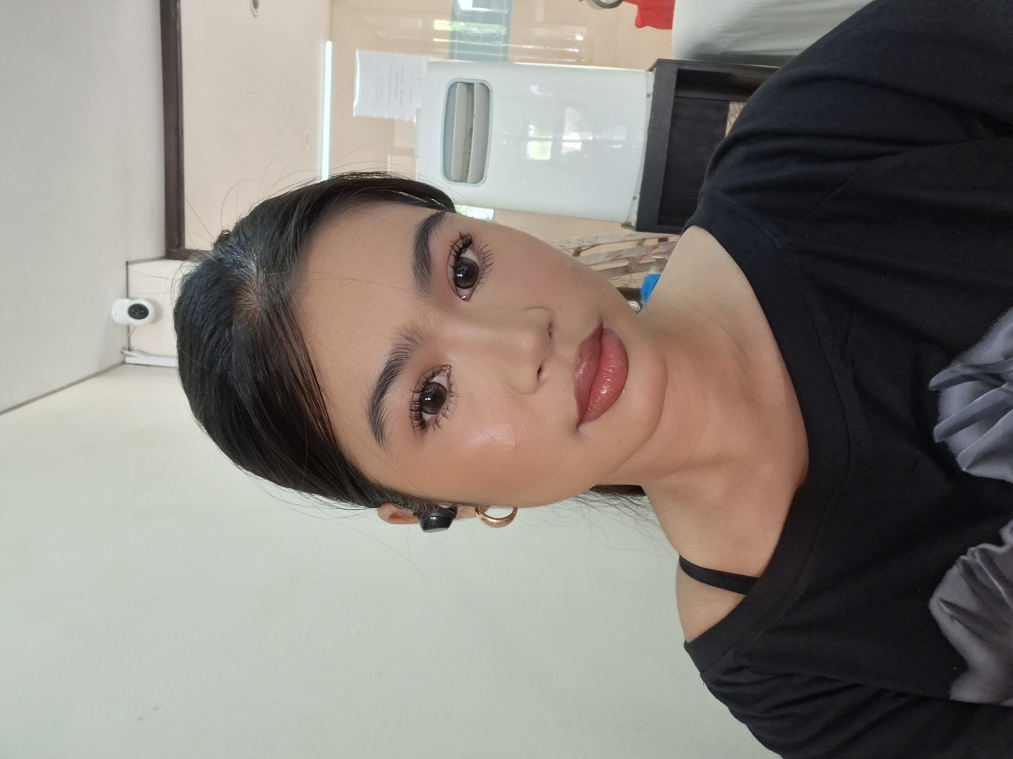 A young woman with dark hair pulled back and wearing makeup, including lipstick and blush, is looking at the camera. She has on a black t-shirt and gold hoop earrings. She is also wearing black earbuds in her right ear. The background shows a white wall, a ceiling-mounted security camera, and a white water cooler with a dark base. The scene appears to be indoors, possibly in an office or waiting area in Kuta, Indonesia.