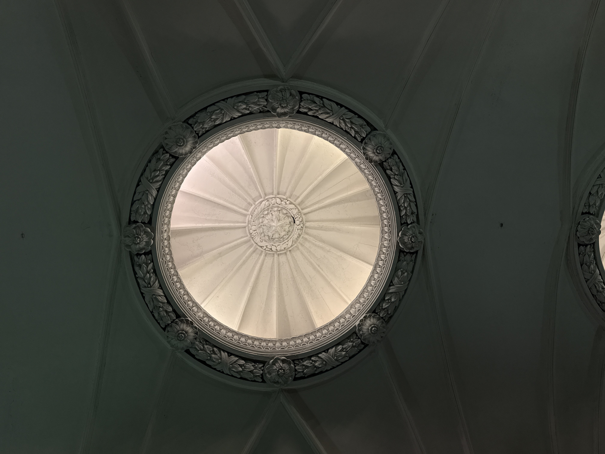 The image shows an interior view of a domed ceiling with a circular skylight, adorned with ornate, classical decorations. The skylight is circular and ribbed, with a decorative medallion at its center. The surrounding frame is embellished with floral and leaf patterns. The architecture is neoclassical, with the ceiling structured into segmented panels. The environment is dimly lit, with the skylight being the primary source of illumination. The location is in Moscow, Russia.