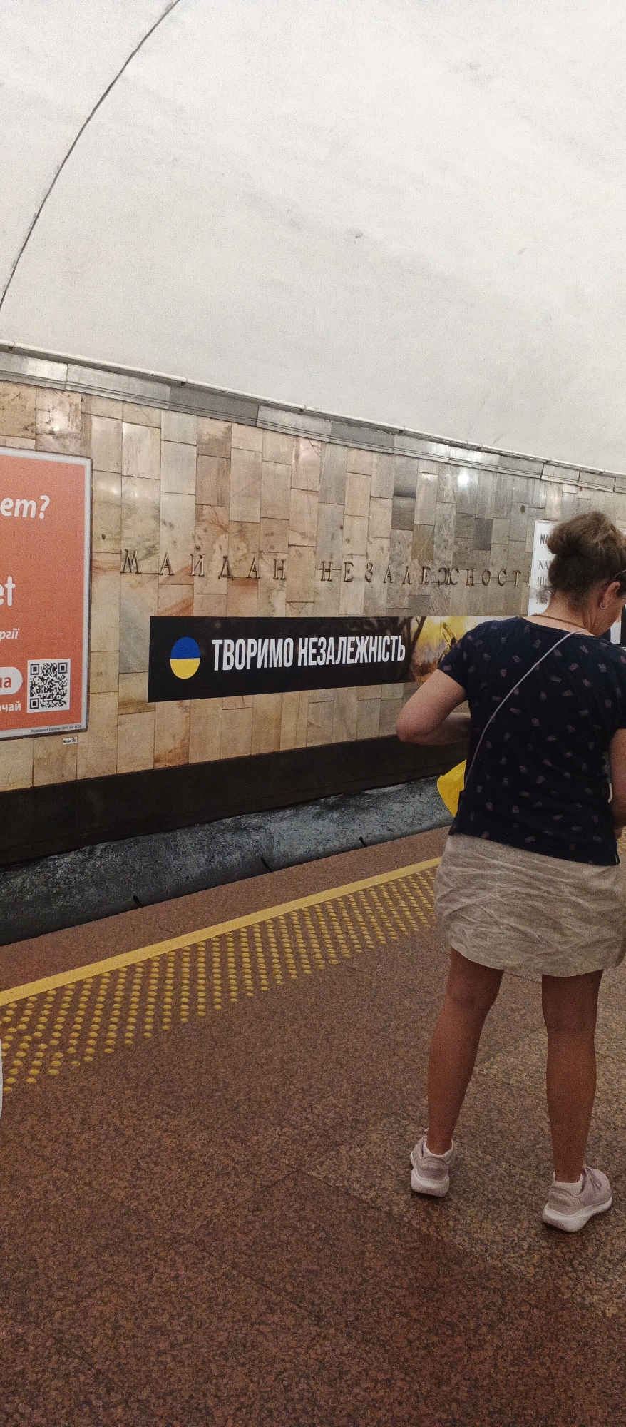 The image shows a section of a subway station, possibly in Ukraine due to the Ukrainian flag and text. The wall of the station is made of marble tiles. There are two signs on the wall. One is a large orange sign with text in an unknown language. The other sign is black and features the Ukrainian flag with the text "ТВОРИМО НЕЗАЛЕЖНІСТЬ" (We are creating independence) in white letters. A woman with her hair in a bun is standing on the platform, facing away from the camera. She is wearing a dark t-shirt with a pattern and a light-colored skirt, and pink sneakers. She is holding a yellow bag. The platform floor is brown with yellow safety markings. In the background, there is a dark trench or gap between the platform and the wall. The ceiling of the station is visible at the top of the image and appears to be a light color with a curved line running across it.