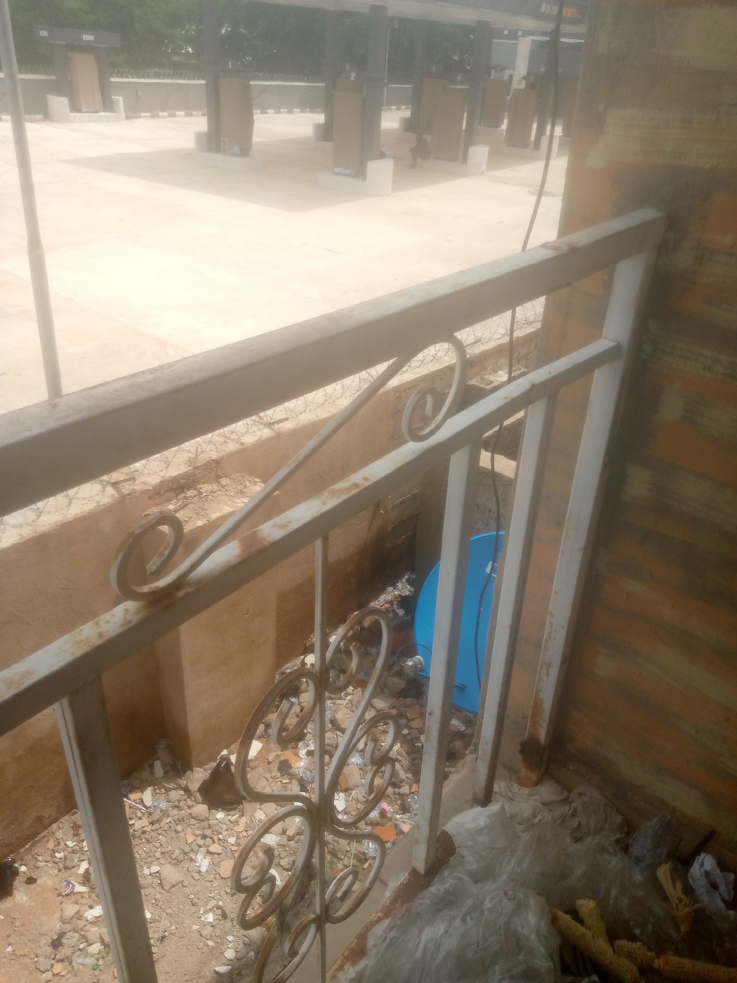 The image depicts an outdoor scene, likely in Jalingo, Nigeria, focusing on a rusty metal railing in the foreground. Behind the railing, there is an area with scattered debris including plastic bags, broken bricks, and other refuse. A bright blue satellite dish is visible amidst the trash. Further in the background, a paved area with several structures that appear to be kiosks or stalls is visible. The structures are rectangular and have doors, with numbers like "1" and "2" painted on some of them. Barbed wire is present along a low wall in the mid-ground, suggesting a security measure. The overall impression is one of an unkempt, possibly commercial or public area.