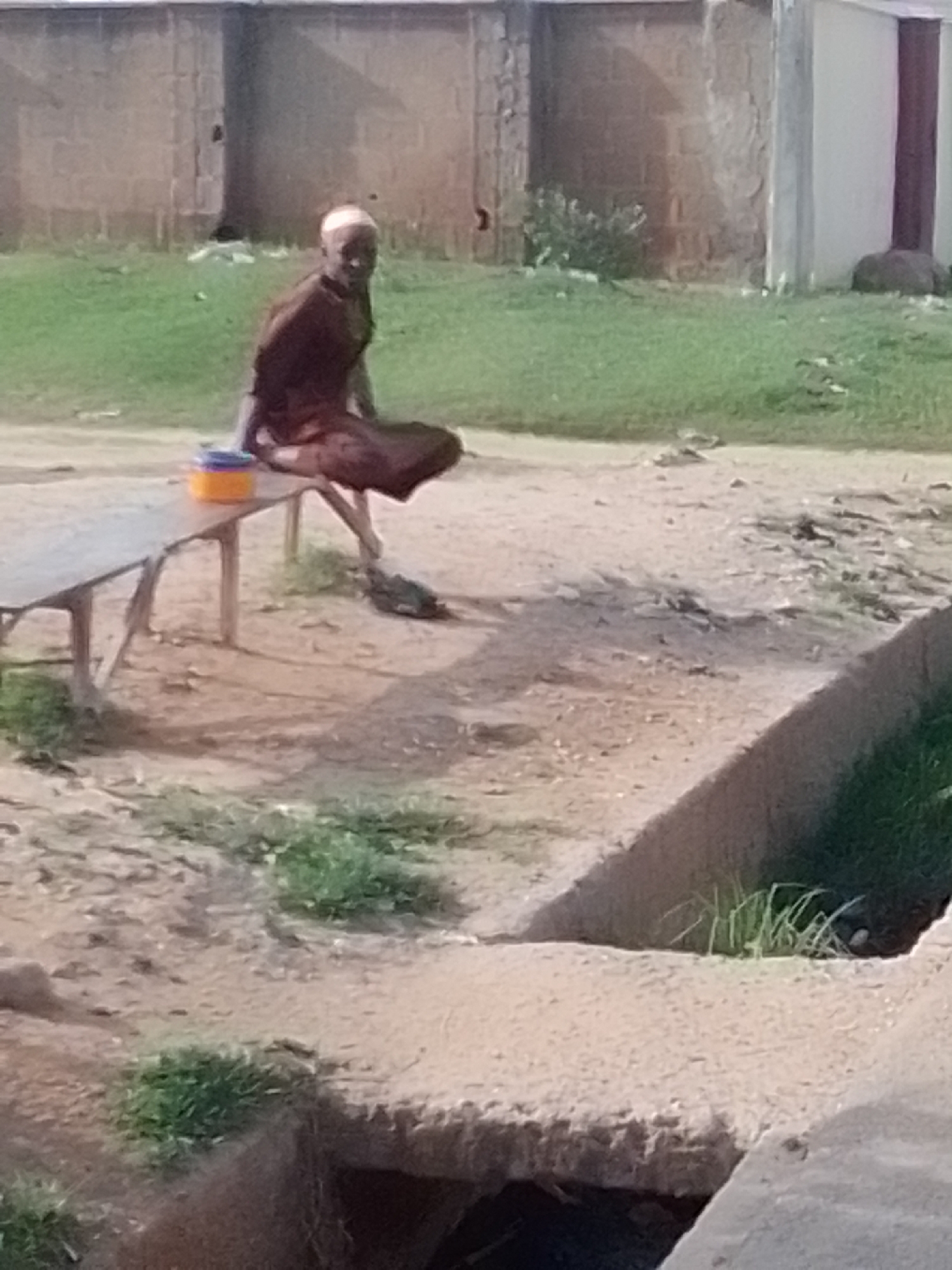 An elderly person, possibly male, is seated on a low wooden bench. They are wearing a dark brown garment and a light-colored cap or headscarf. Beside them on the bench is a blue and orange container. The person is facing slightly towards the camera, with their legs crossed. The bench appears to be outdoors, on a dirt or sandy area with patches of green grass. In the background, there is a brick wall and a white building. A drainage ditch runs along the right side of the foreground. The scene suggests a public or semi-public outdoor space, possibly in a less developed area of Gombe city, Nigeria, given the presence of the brick wall, building construction, and natural drainage channel. The lighting suggests it is daytime.