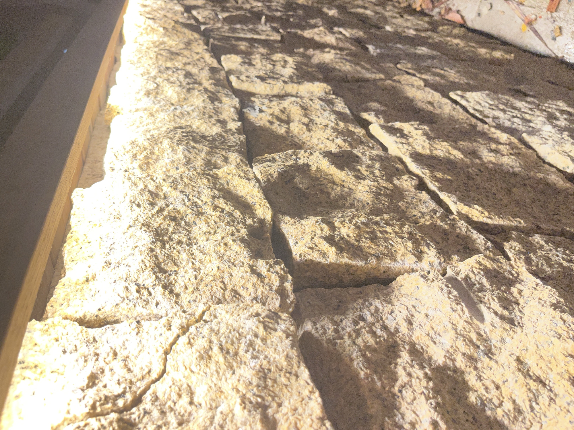 The image shows a close-up view of a wall constructed of tan-colored stone blocks. The blocks are arranged in a staggered pattern, with visible joints between them. The surface of the stones is rough and textured. The lighting in the image is uneven, with bright light illuminating the upper left corner and casting shadows on the right. The background is slightly out of focus, showing some debris. A portion of a wooden frame is visible on the left edge of the image. The location is Bac Nha Trang Ward, Vietnam.