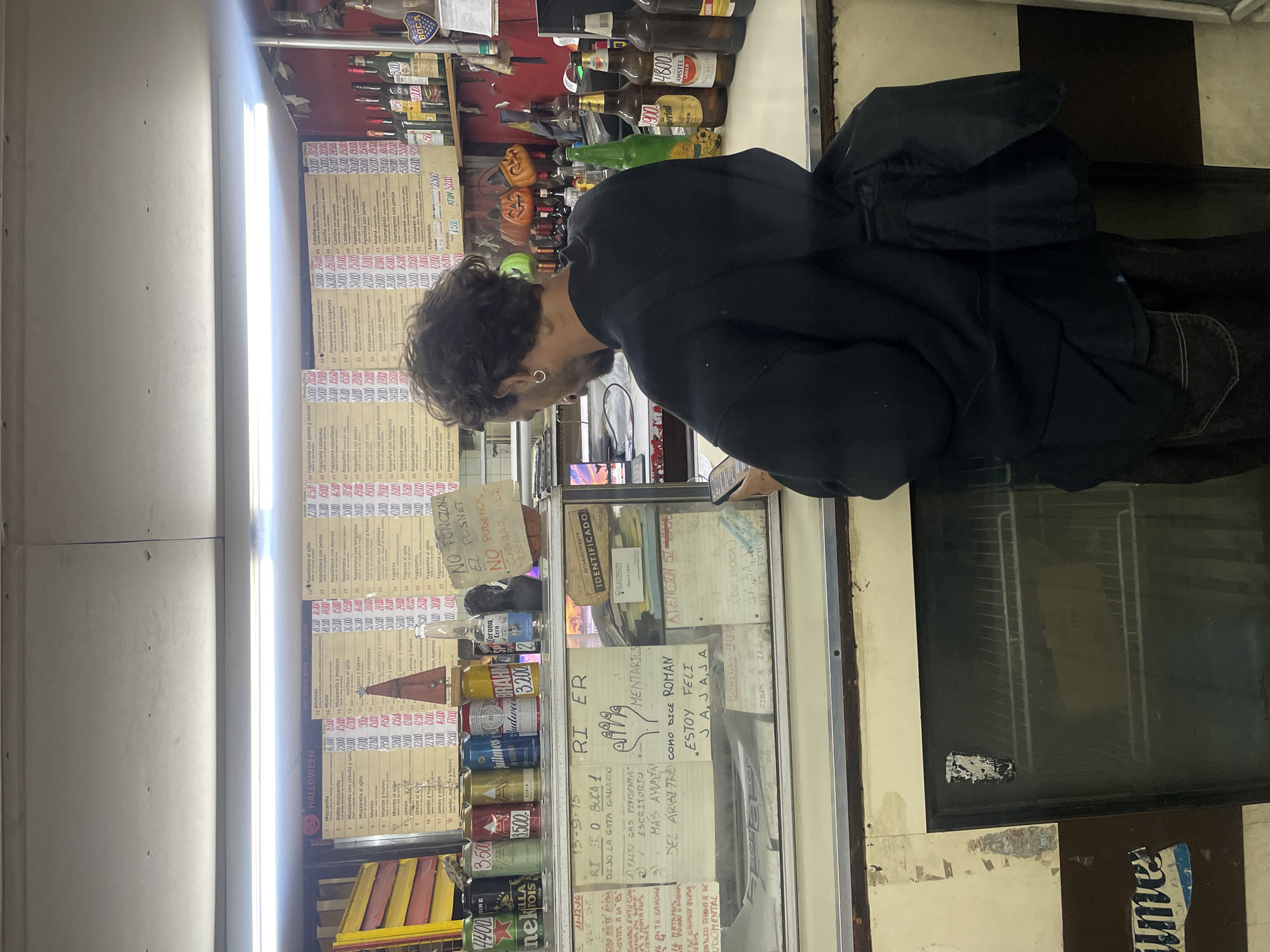 A man stands in front of a storefront in Vicente López, Argentina. He is looking at a handwritten menu and a glass display case filled with canned goods and price lists. The sign on the case has handwritten notes that include "RI ER O BUCA 1," "Falto Gas PIFICENTA", and "ESTOY FELI JA JAJA". The menu board on the left lists various pizza toppings and prices. The store also sells beer and other drinks. The storefront has a window that is lit by a fluorescent light. A refrigerator with a glass door is visible on the right. A partially visible street sign says "aume". The location is most likely in the province of Buenos Aires, Argentina.