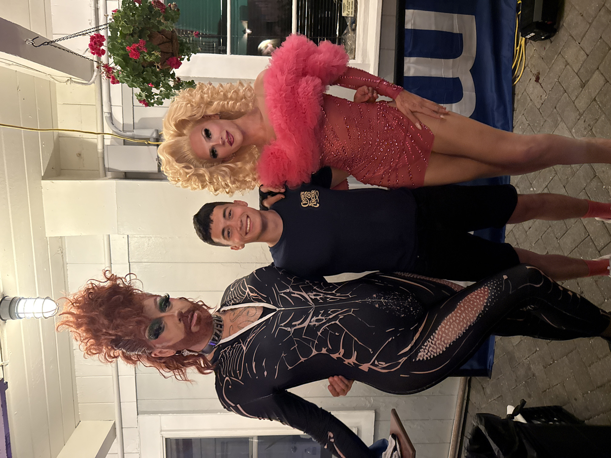 In an outdoor setting in Provincetown, Massachusetts, two drag performers and a civilian stand together. The performer on the left, with voluminous reddish-brown hair styled into a mohawk, wears a black full-body suit with a spiderweb-like design in light pink. They also sport a choker necklace and heavy eye makeup. The performer on the right, with blonde curly hair, is dressed in a form-fitting pink sequined dress with a ruffled neckline and sleeves. The civilian, a young man in a navy blue t-shirt and black shorts, stands between the two performers with his arms around them. The background includes a white building with a light fixture, hanging plants with red flowers, a window, and a blue banner. The ground is paved with cobblestones.