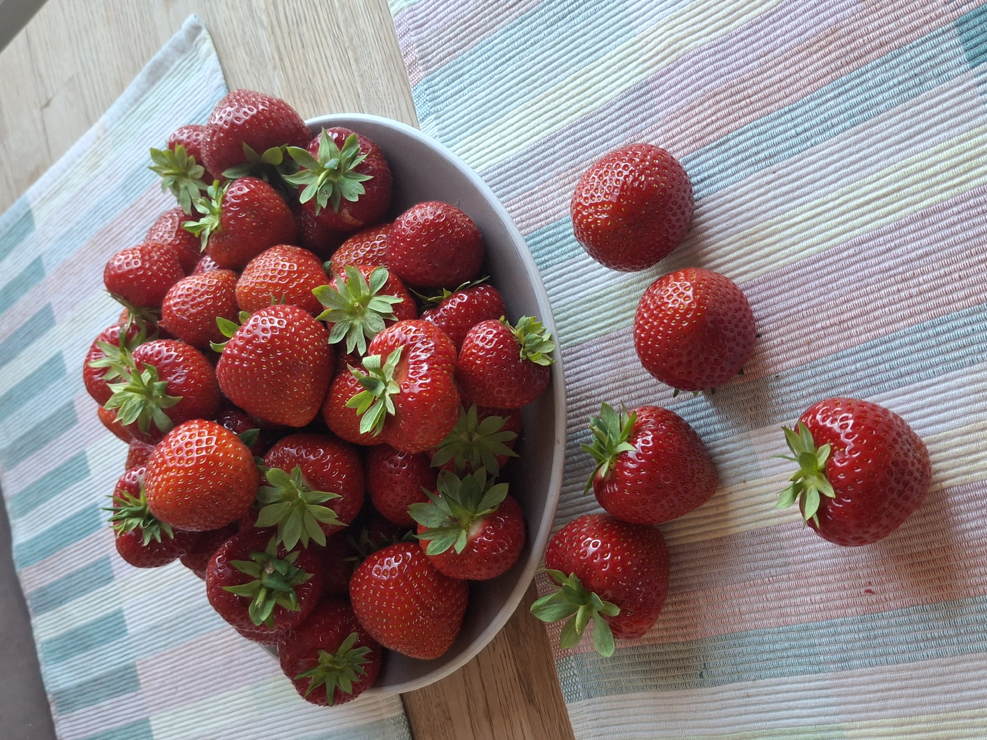 A bowl of fresh strawberries sits on a table. The bowl is filled with strawberries, some of which are stacked on top of each other. Several strawberries are also scattered around the bowl on a striped placemat. The strawberries are bright red and appear to be ripe. The background is a light-colored wooden table. This image was taken in Åsgårdstrand, Norway.