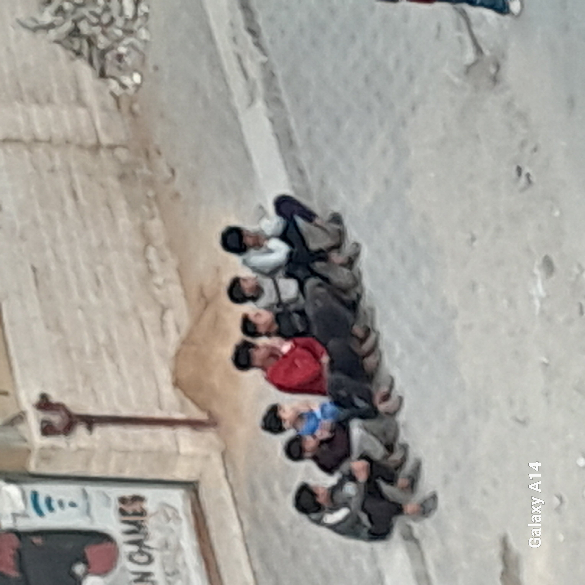 The image captures a group of approximately eight to nine individuals, appearing to be young males or boys, lying in a line on a light-colored ground surface, likely a pavement or street. They are positioned close to each other, with their bodies generally oriented in the same direction.
The subjects are dressed in casual clothing, including a red top, a blue top, a white shirt, and various dark jackets and trousers. Their faces are mostly obscured or out of focus due to the image's resolution and perspective. The activity appears to be one of resting or waiting, as they are all reclined on the ground.
The setting is outdoors during the daytime, suggested by the bright, even lighting, although specific weather conditions are not clear. To the left of the group, a light-colored, textured wall or building facade is visible. In the bottom left corner, a portion of a sign or poster is visible, displaying the text "...EN GAMES". The ground itself has a somewhat rough or patterned texture.
The image is captured from an elevated and tilted perspective, which contributes to the somewhat blurry and pixelated quality, especially when examining finer details. The text "Galaxy A14" is prominently displayed vertically along the right side of the image, indicating the device used to take the photograph. No specific geographical location cues are evident beyond a general urban or semi-urban environment.