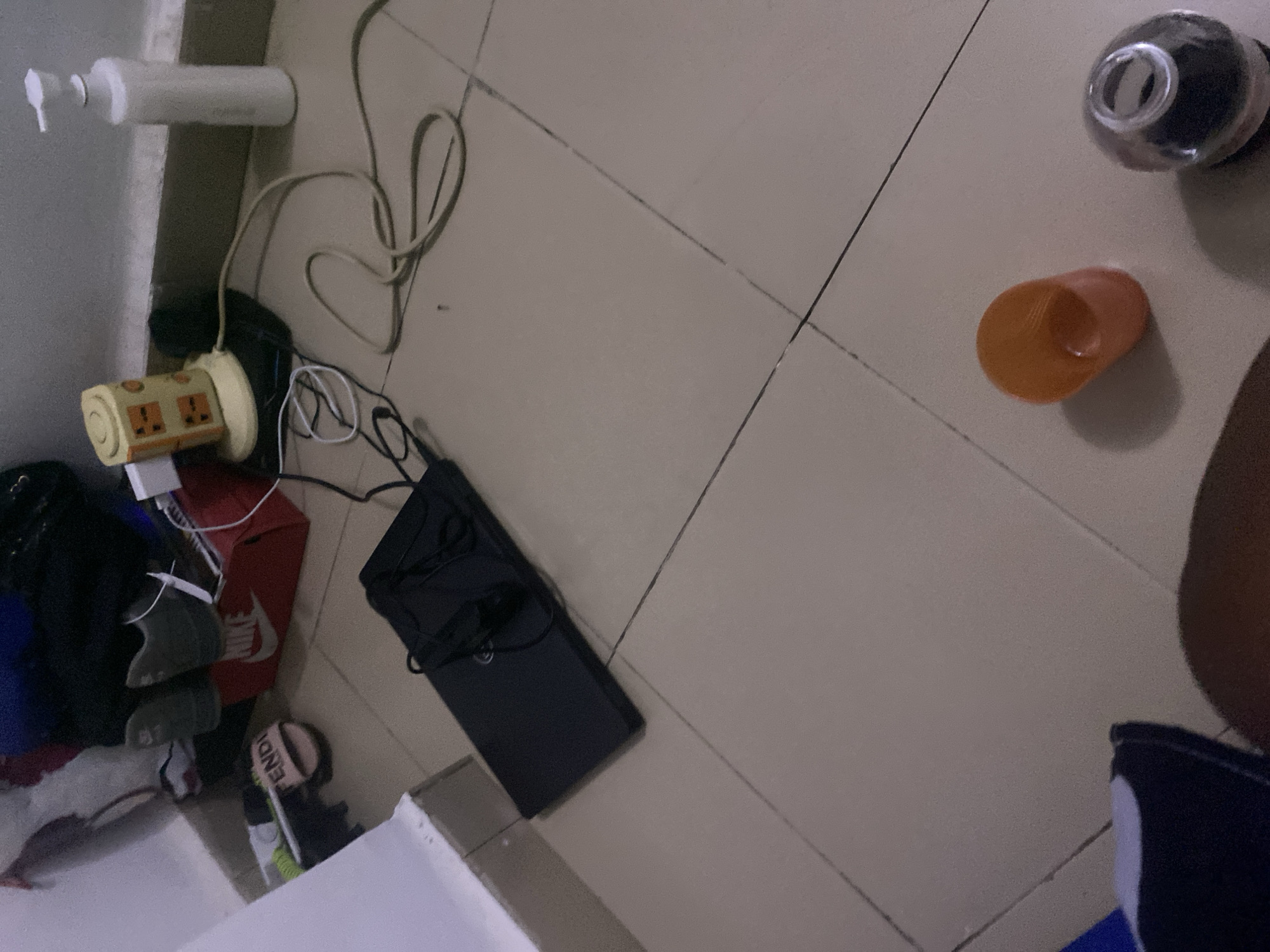The image depicts a tiled floor in a room, possibly a bedroom, in Howayi, Nigeria. Various objects are scattered on the floor, including a black laptop, a multi-outlet power strip with cables, a white pump bottle, a red Nike shoebox containing two pairs of sneakers, a pink Fendi slipper, an orange plastic cup, and a clear glass container with a dark substance inside. The overall scene suggests a cluttered living space.