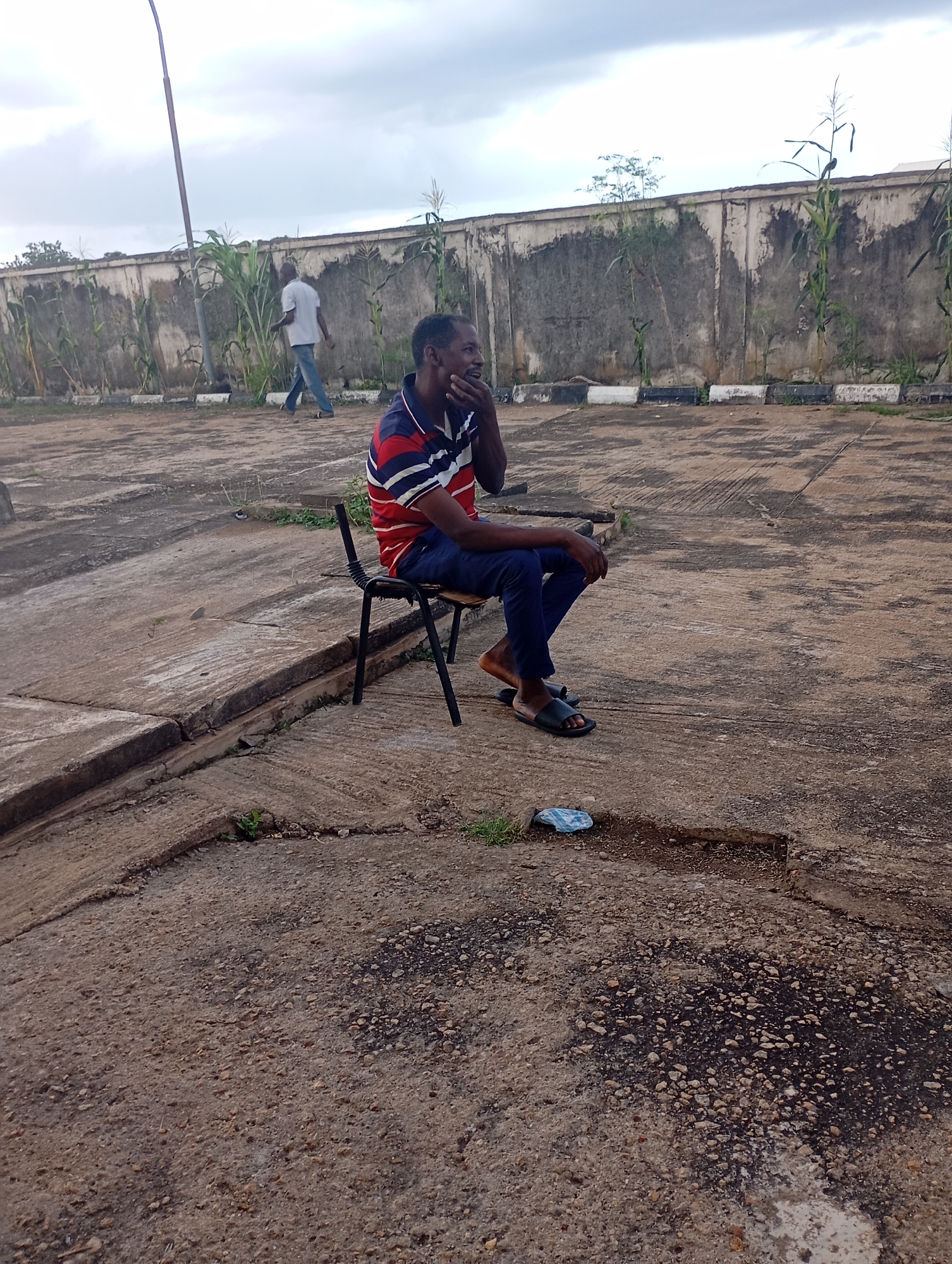 A man in a red, white, and blue striped shirt and dark pants sits on a black metal chair outdoors. He is looking to the side and has his hand near his mouth. He is wearing dark sandals. In the background, a concrete wall with some plants growing near it separates the paved area from a cloudy sky. Another man in a light-colored shirt and blue jeans walks away from the camera in the background, in front of the wall. The ground is made of cracked and uneven concrete. A blue plastic bag lies on the ground near a pothole. The scene appears to be an outdoor area in Shendam, Nigeria, possibly a parking lot or courtyard.