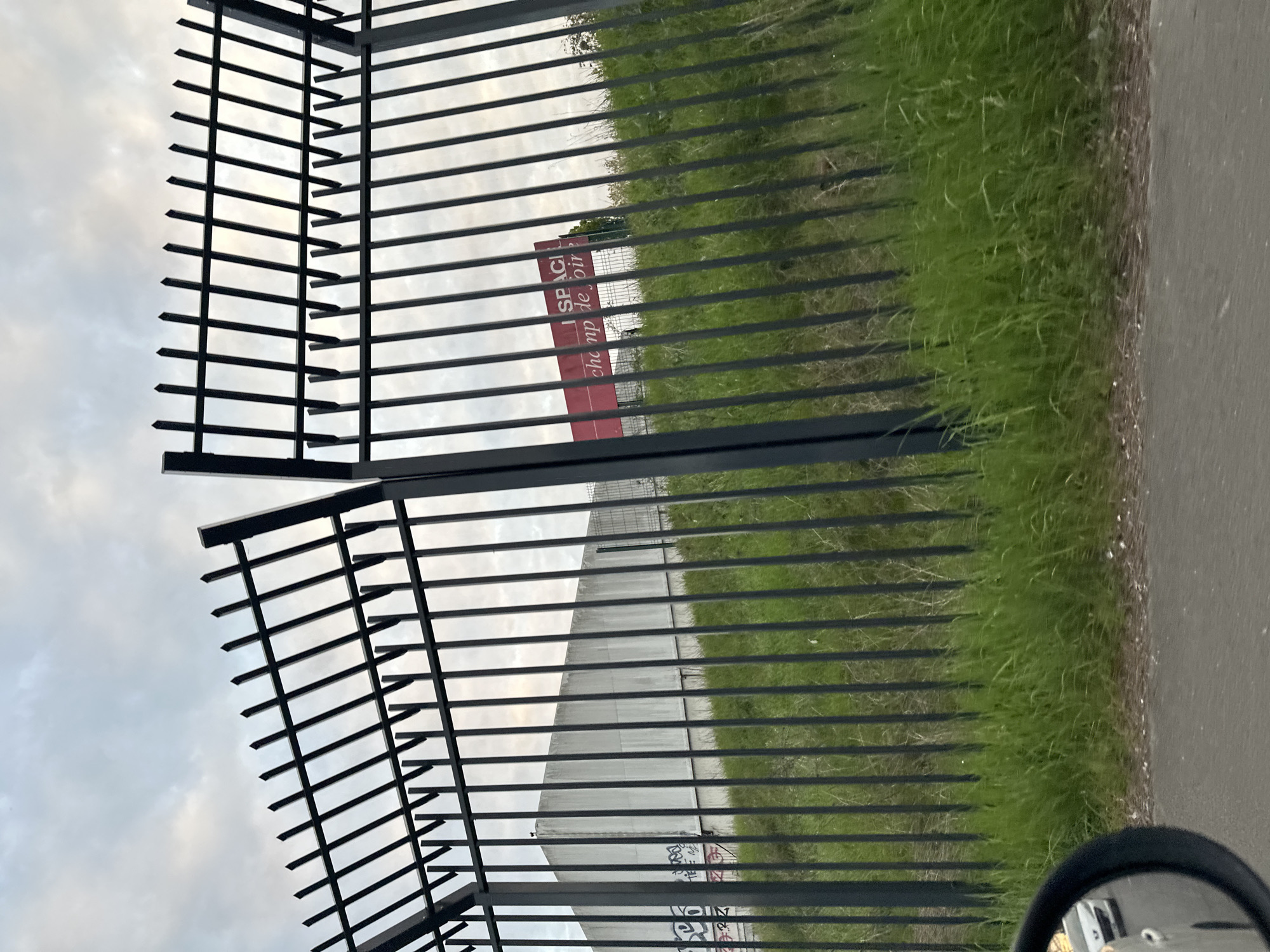 The image shows a black metal fence. Beyond the fence is green grass and a road. A sign is visible through the fence with the words "ESPACE champ de foire". Graffiti is visible on a concrete structure. The location is likely Sarcelles, France. A side mirror is visible in the bottom right. The sky is cloudy.