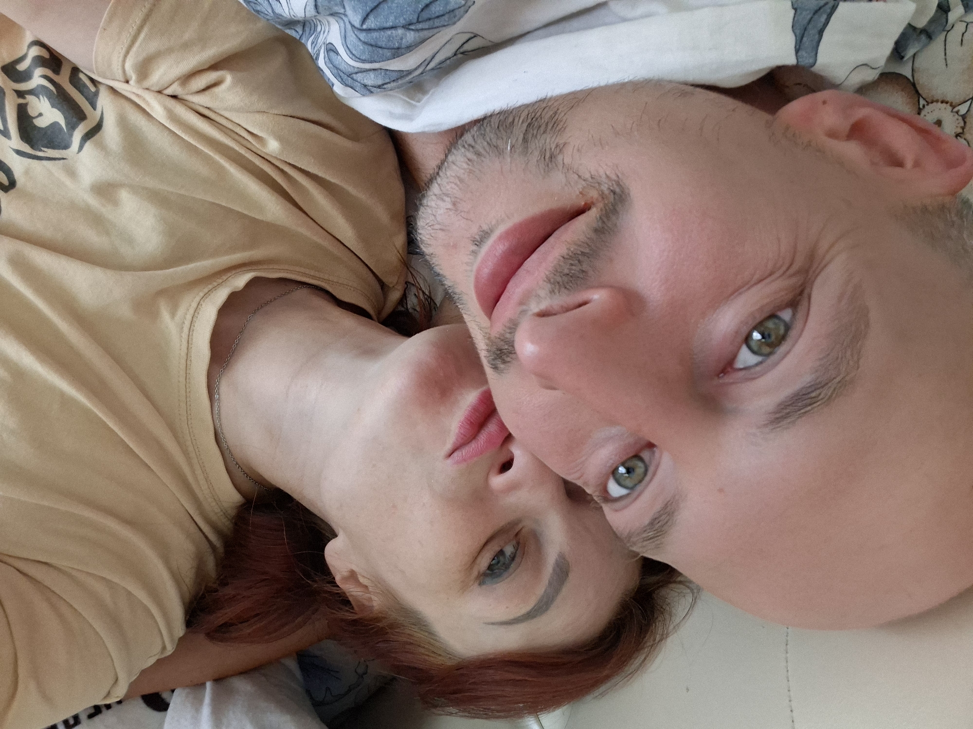 The image is a close-up selfie of a man and a woman lying down, possibly in bed. The man is on the left, with his face close to the camera, showing his green eyes and a partial smile. He has a shaved head and stubble. The woman is on the right, her head resting near the man's. She has red hair and her lips are pursed as if for a kiss. She is wearing a tan t-shirt and a silver necklace. The background appears to be a bed with patterned sheets on the left and a white surface on the right. There is no visible indication of the location being in the city of Myadzyel, Belarus.