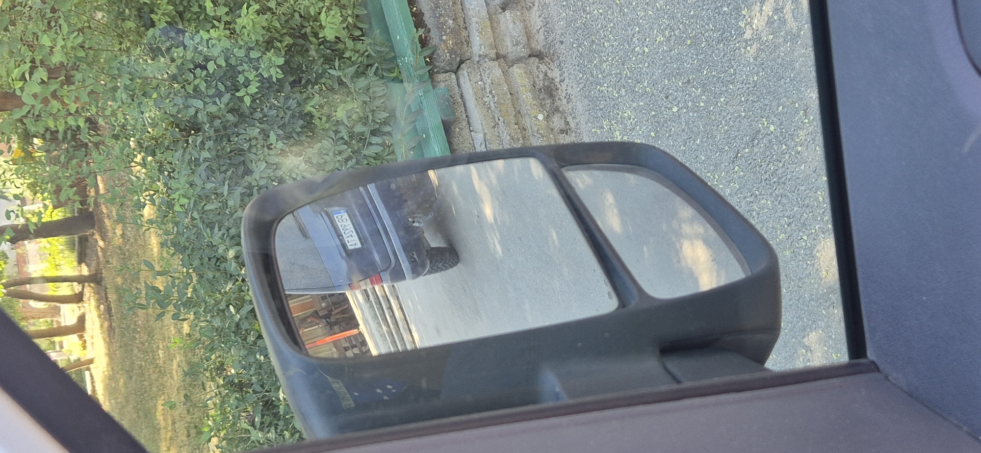 In a side-view mirror of a vehicle, the rear of a dark blue minivan is visible. The minivan has a license plate that reads "AT ASFF 89". The minivan is parked on a paved surface. Behind the minivan, there are some stone steps leading to a building with a red facade. In the background, there are trees and green foliage. The scene appears to be in an outdoor environment, possibly a residential area or a park.