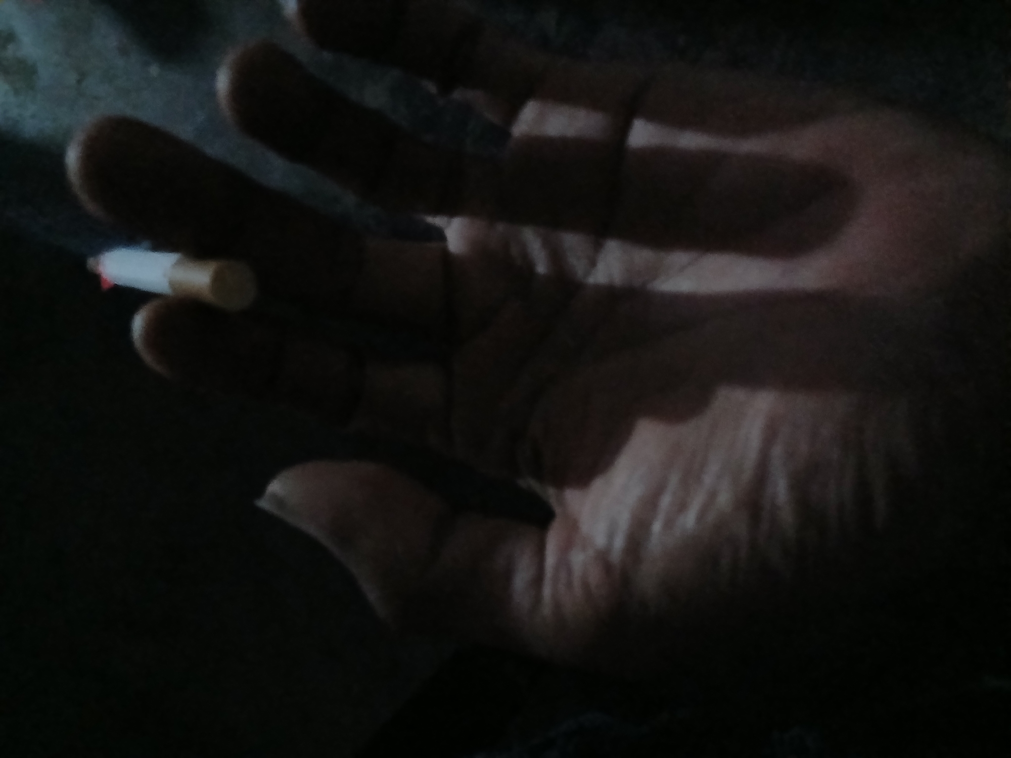 A close-up, dimly lit image shows a hand holding a lit cigarette. The cigarette is positioned between the thumb and index finger, with the glowing end visible. The hand is in the foreground, with its palm facing upwards, and the texture of the skin, including wrinkles and creases, is discernible despite the low light. The background is dark and out of focus, making it difficult to discern specific environmental details. The overall impression is that of a personal moment captured in a low-light setting. The image appears to be a photograph, possibly taken by a mobile phone given the lighting and composition.